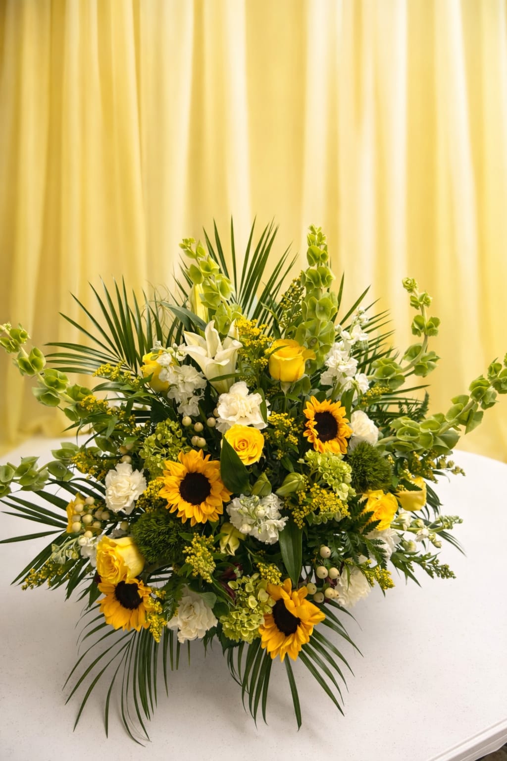 Morning sunshine - Bright yellow sunflowers and roses take center stage in this large celebratory floral arrangement, making it an ideal flower arrangement for birthdays, congratulations, or as an uplifting sympathy tribute. The strong vertical lines and palm-like fronds create a dramatic silhouette that reads both joyful and respectful-perfect when you want the person you're thinking of to feel genuinely seen and celebrated.  This full design pairs sunny yellow blooms with crisp white chrysanthemums, roses, and carnations, set against abundant green accents for a fresh, layered look. Tall vertical elements rise above a rounded base, giving the piece impressive height and presence; palm-style foliage extends outward for added drama and texture. The overall palette of yellow, white, and green keeps the impression bright and hopeful while maintaining a formal, structured composition.  Because of its scale and bold presence, Morning sunshine works best as a statement piece for a room, entryway, or memorial setting where it can be seen and enjoyed from a distance. Standard care: keep water topped up, trim stems every few days, and place away from direct heat to extend bloom life.   If any design elements are of major importance to your order, please include them in the florist instructions at checkout or contact us to ensure availability.
