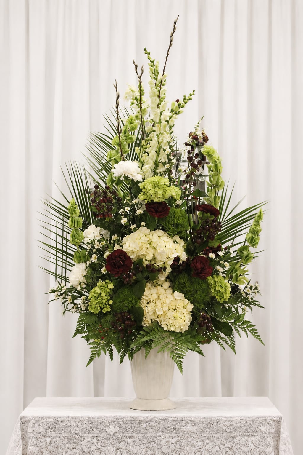 Eternal grace - Tall, layered white hydrangeas and architectural greenery make an immediate, comforting impression-this sympathy arrangement is designed to convey quiet respect and heartfelt remembrance. The formal, ceremonial silhouette in a white ceramic pedestal vase gives it presence on a memorial table or at a funeral service, the deep burgundy accents lending a note of solemn warmth.  Clusters of lush white hydrangeas form the body, complemented by green blooms, carnations and snapdragons that add vertical movement; palm fronds and fern-like foliage fan outward to create an elevated, sculptural profile. Long vertical stems rise above the main mass for added height and dignity, and the ceramic pedestal anchors the piece with classical elegance. Sized for a table or display, it reads as a large, event-style arrangement meant to fill a space and set a reverent tone.  A meaningful choice when you need to honor someone's memory or mark a solemn event, it offers the kind of presence that helps mourners feel seen and supported. For placement on memorial tables or service platforms; please note the arrangement is designed for display rather than hand-carried use and should be kept in a cool area away from direct heat to preserve bloom life.   If any design elements are of major importance to your order, please include them in the florist instructions at checkout or contact us to ensure availability.
