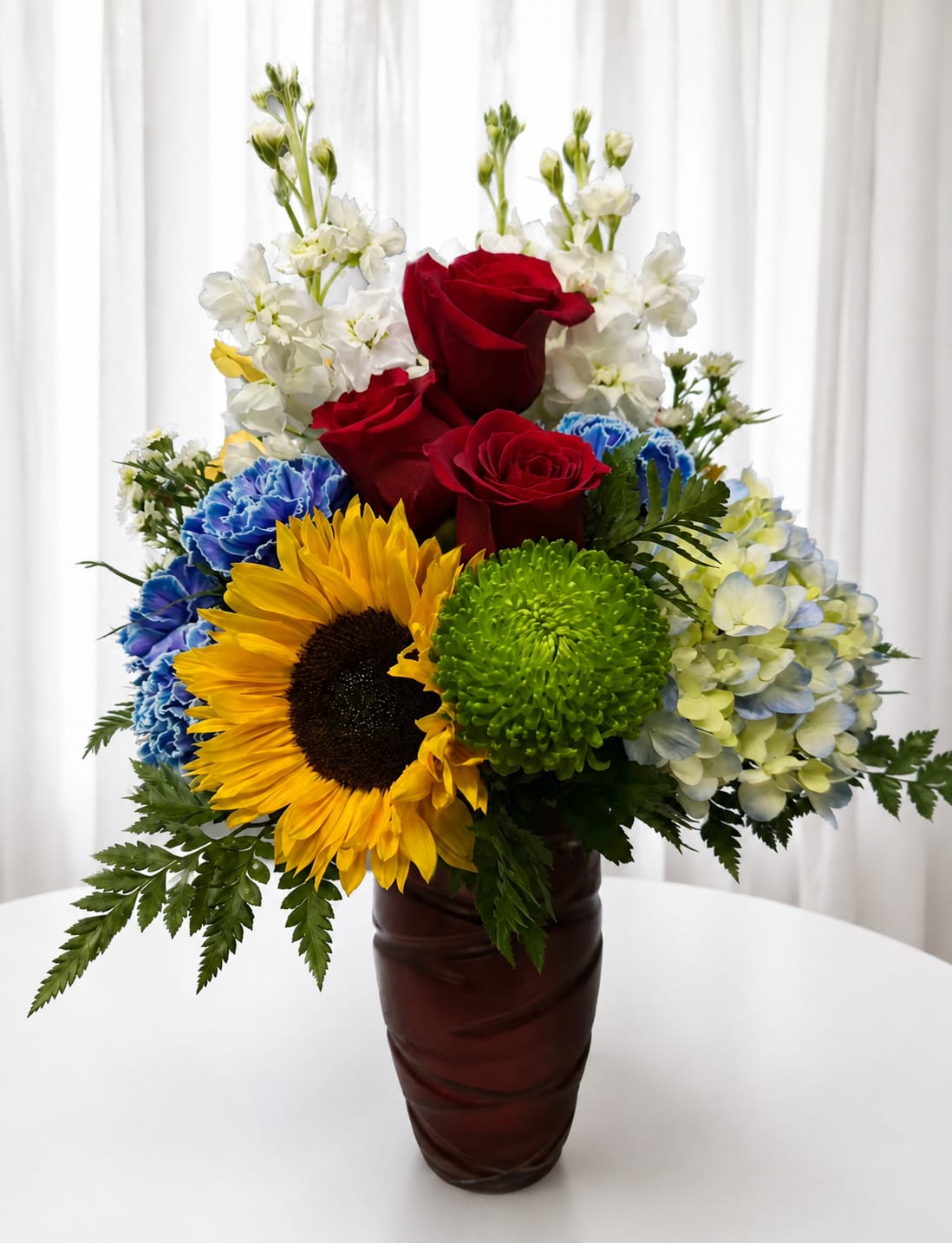 Patriotic Sunshine - A bright sunflower center surrounded by deep red roses and bold blue carnations makes an immediate, celebratory impression - perfect as a patriotic birthday bouquet, congratulations arrangement, or simply to brighten someone's day. This flower arrangement for everyday gifting combines the warmth of sunlit yellow and the drama of true red and blue to create a joyful, confident statement your recipient will notice the moment it arrives.  The composition pairs a large yellow sunflower with clustered deep red roses, blue carnations, white stock-like stems for airy height, and a green chrysanthemum focal accent, arranged in a tall ceramic vase. Blooms are layered with larger flowers grouped lower and lighter white stems rising above for movement, giving the arrangement a full, textured silhouette that reads lively from every angle.  Send this to someone you want to celebrate - a graduate, a neighbor, or a friend marking a milestone - and they'll feel noticed and cheered. The tall ceramic vase provides immediate display readiness; to keep blooms looking their best, refresh the water every other day and trim stems as needed for longer enjoyment.