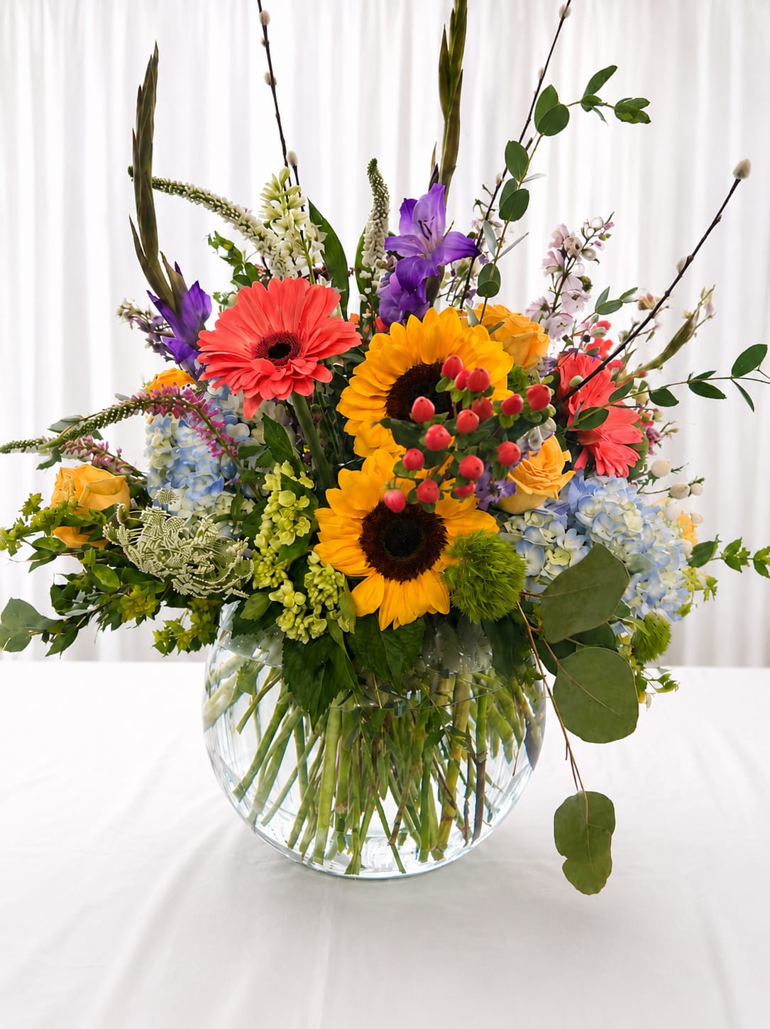 Something pretty  - A burst of sunny joy-this mixed flower bouquet brings together bold sunflowers, cheerful yellow roses, pink gerbera-style blooms, blue hydrangea, and purple accents to create a vibrant birthday bouquet or celebratory arrangement that instantly lifts the room. The lively, garden-inspired silhouette-tall branching stems and berry-like accents rising above the main body-feels spontaneous and joyful, the kind of gift that makes the person you're thinking of feel truly celebrated.  The flowers are gathered in a clear round glass vase so the stems show below the waterline, giving a clean, contemporary finish that highlights the arrangement's lively colors: bright yellows, soft pinks, cool blues, and pops of purple. Textures range from the sunflowers' bold faces to the hydrangea's cloudlike clusters and the delicate, airy accents, creating visual depth and movement.  Perfect for birthdays, congratulations, or when you simply want to brighten someone's day, this bouquet suits recipients who love bold color and a natural, garden-fresh look. The clear vase makes care simple-fresh water and a clean vase keep stems healthy-so your gift continues to feel like a sunny celebration for days.   If any design elements are of major importance to your order, please include them in the florist instructions at checkout or contact us to ensure availability.