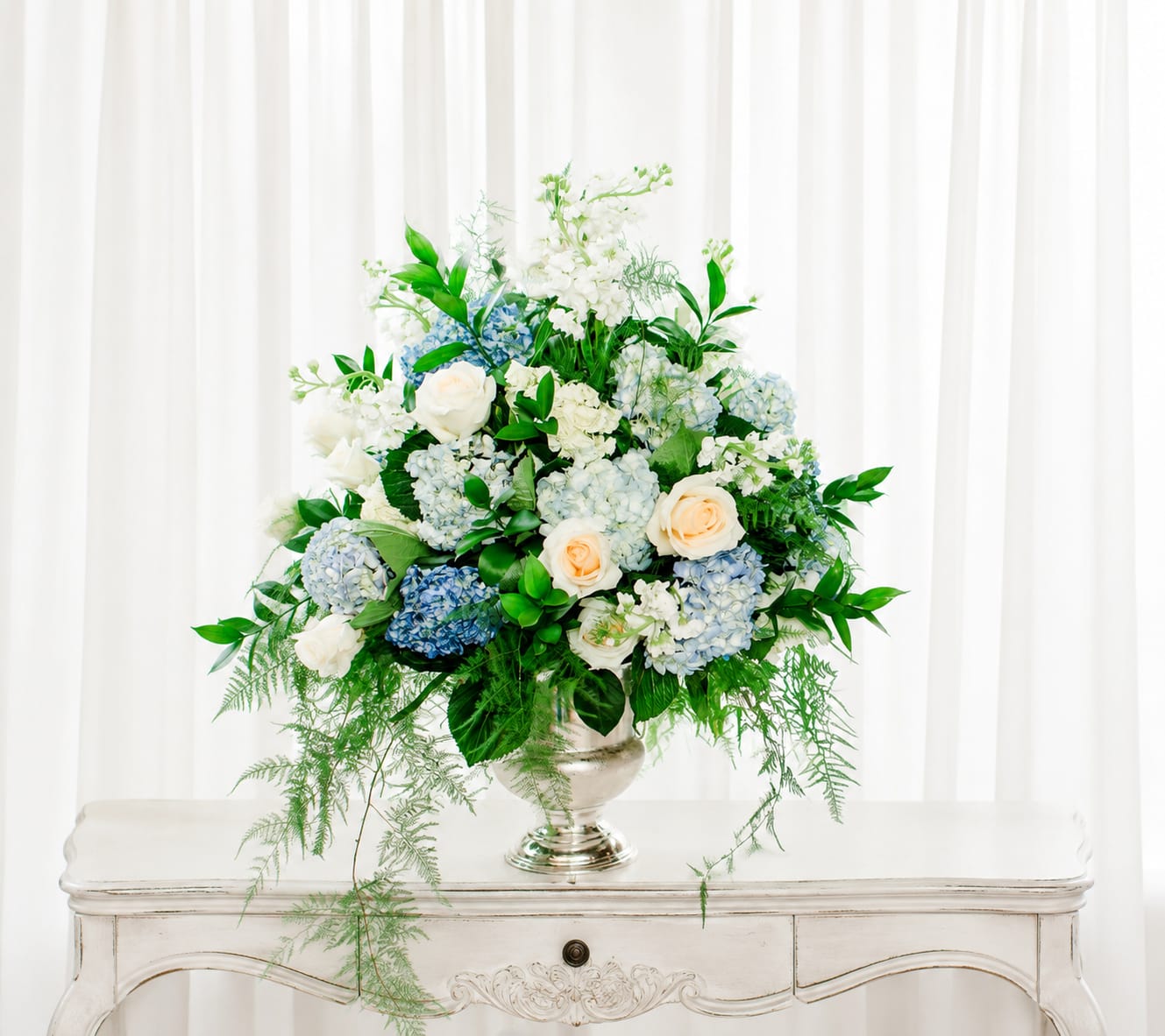 Special moments - A lush, rounded design of white and pale blue hydrangeas and cream roses creates an elegant statement bouquet perfect for a wedding bouquet display, sympathy arrangement, or refined event centerpiece. The cool palette of white, blue, green, and soft peach accents reads fresh and calming-the kind of arrangement that makes the person you're thinking of feel comforted and noticed on an important day.  This arrangement pairs full, cloud-like hydrangeas with classic cream roses and small white accent blooms, woven through with bright green foliage and delicate trailing fern-like stems for gentle movement. Presented in a silver pedestal vase, the composition has a formal yet airy presence that suits tabletops, altars, or sideboards; the silver adds a timeless, polished touch that complements the soft colors without overpowering them.  Ideal for marking weddings, memorials, anniversaries, or any moment that calls for refined sympathy or celebration, this arrangement arrives ready to display in the included silver vase. The generous rounded size fills a medium to large space-ask us about delivery timing to ensure peak freshness for the occasion.   If any design elements are of major importance to your order, please include them in the florist instructions at checkout or contact us to ensure availability.