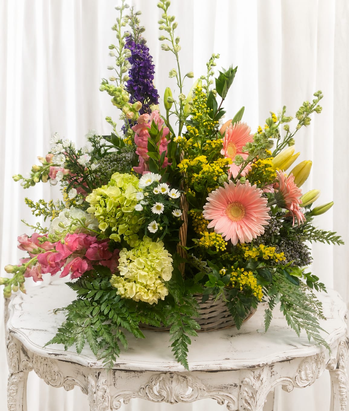 Spring time - A lively basket of springtime joy, this mixed flower arrangement is the perfect spring gift or cheerful birthday bouquet to make someone feel truly celebrated. Pink gerbera daisies and tall purple snapdragon-like spikes create an uplifting vertical rhythm while pale green hydrangea clusters and delicate white daisies fill the base, giving the person you're thinking of a fresh, garden-picked impression the moment it arrives.  Arranged in a rustic wicker basket, the composition blends pastel pinks and purples with bright yellow filler blooms and ferny greenery for a loose, airy silhouette that's rich in texture. Tall stems rise above a fuller, abundant base so the design reads both generous and casual - an ideal centerpiece for a brunch table or a sunny windowsill. The mix of gerbera daisies, hydrangeas, snapdragon-like spikes, small daisies, and additional seasonal blooms offers varied shapes and soft scents that feel like a spring stroll.  Perfect for celebrating birthdays, new beginnings, or simply saying "I'm thinking of you," this basket sends a warm, uplifting message to its recipient. Order with confidence for local delivery; the wicker container keeps the arrangement easy to display and enjoy, and simple care-fresh water and a cool spot away from direct heat-will help the flowers look their best for days.   If any design elements are of major importance to your order, please include them in the florist instructions at checkout or contact us to ensure availability.