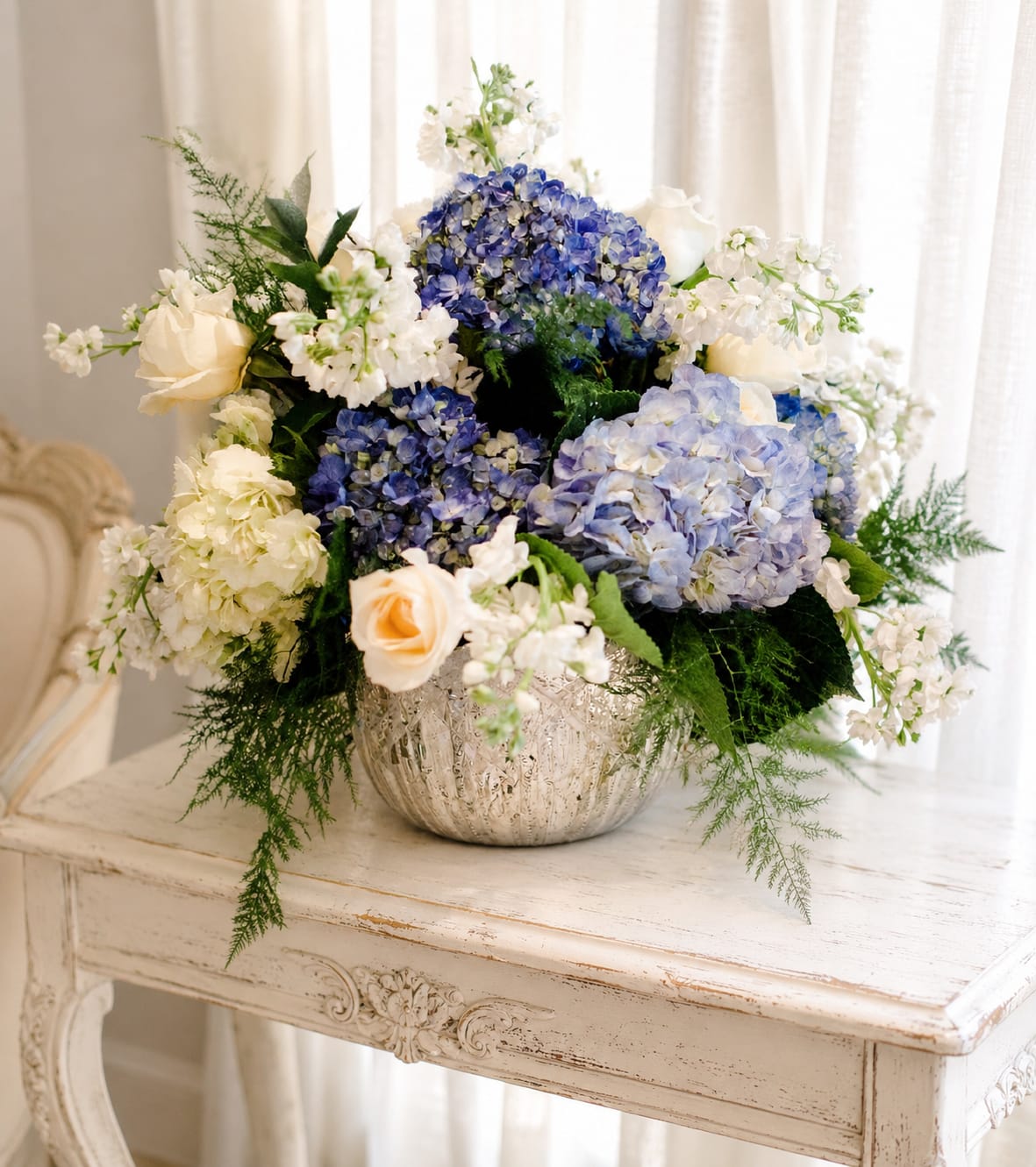 Magic moments - Big, cloud-like clusters of blue and pale blue hydrangeas set the tone for this elegant flower arrangement - a refined wedding centerpiece or anniversary bouquet that reads as romantic and serene. The rounded, garden-style composition pairs those hydrangeas with crisp white blooms and a few soft peach roses so the person you're celebrating will feel both comforted and cherished in equal measure.  Textured, feathery greenery and airy white filler flowers extend outward from the focal blooms for depth and movement, creating a full, layered look. Arranged in an ornate silver vase and photographed on a distressed white wooden table, the palette of blue, white, green, and peach reads both classic and fresh; this makes it equally at home on a reception table, mantel, or as a meaningful gift to mark a special moment.  Perfect for weddings, anniversaries, or a thoughtful "I'm thinking of you," this design is crafted to make the recipient feel truly seen. Ask about available sizes or vase options when ordering to match your delivery needs and placement - light trimming of stems and fresh water will keep those lush hydrangeas and roses looking their best.   If any design elements are of major importance to your order, please include them in the florist instructions at checkout or contact us to ensure availability.