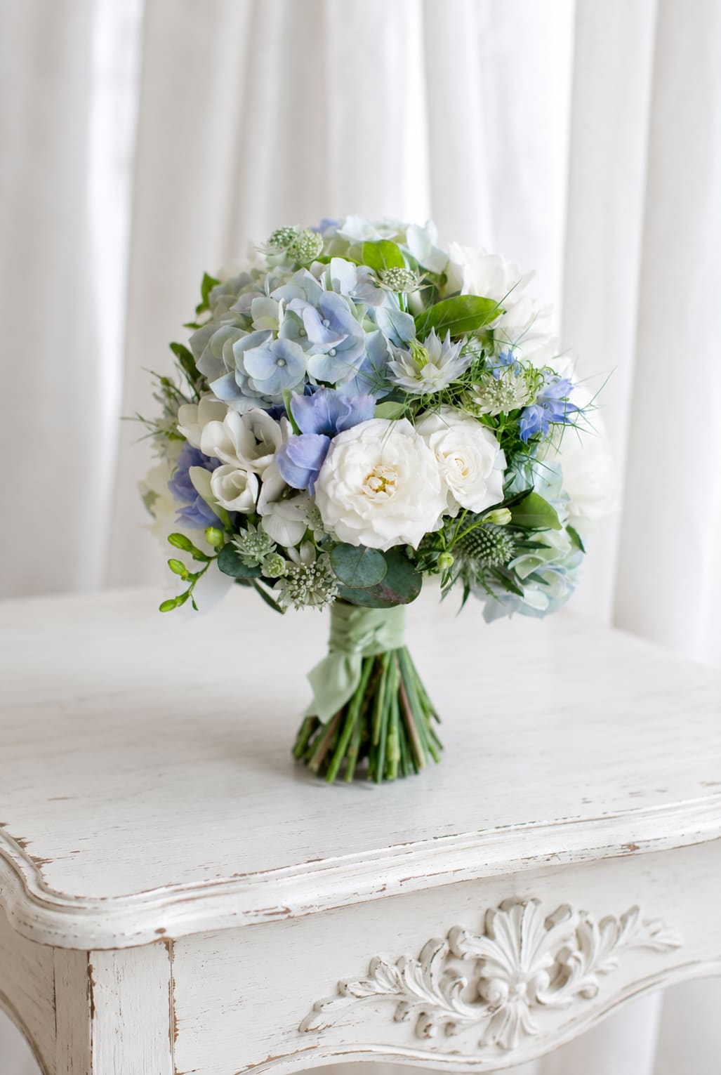 Something blue - A soft, romantic bridal bouquet in white, pale blue, and touches of green, this hand-tied arrangement is perfect as a wedding bouquet or for a formal celebration where understated elegance matters. The rounded, bridal-style shape feels classic and refined - the kind of bouquet that will make the person carrying it look and feel gracefully celebrated on their big day.  Large hydrangea clusters set the airy foundation, joined by white rose-like blooms and delicate ranunculus accents for a mix of lush volume and gentle texture. Smaller filler flowers and fresh green touches soften the palette and add movement, while the stems are gathered tightly and finished in a pale green satin ribbon for a polished, comfortable grip.  Ideal for brides, bridesmaids, or as a centerpiece for an intimate ceremony, this bouquet reads as timeless and effortlessly pretty. To keep the blooms looking their best, handle gently, keep stems in water until the moment they're needed, and store in a cool place away from direct sunlight before use.   If any design elements are of major importance to your order, please include them in the florist instructions at checkout or contact us to ensure availability.