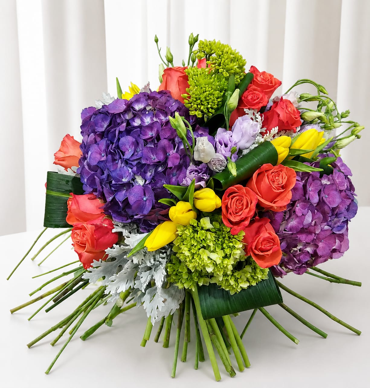 Bold and beautiful wedding bouquet - A dramatic cluster of large purple hydrangea heads anchors this wedding bouquet, juxtaposed with coral-red roses and bright yellow tulips to create a vivid, celebratory palette perfect for a wedding, anniversary, or joyful gift. The mix of bold purples, warm reds, and sunny yellows reads festive and romantic-an arrangement that makes the person you're thinking of feel truly celebrated the moment they see it.  This arrangement is densely packed for a lush, full look: oversized hydrangea blooms, classic roses, tulips, and small green clustered filler blooms combine for rich texture and depth. Silver-toned foliage adds a cool metallic contrast while a broad decorative leaf wrap provides structured support and a polished finish, giving the bouquet a modern, sophisticated silhouette.  Ideal for brides seeking a colorful statement bouquet or for sending as a standout birthday or anniversary gift, this design reads joyful and heartfelt. Arrange for same-week delivery when you need it fresh; to keep blooms vibrant, recut stems and place in clean water upon arrival and change water daily.   If any design elements are of major importance to your order, please include them in the florist instructions at checkout or contact us to ensure availability.