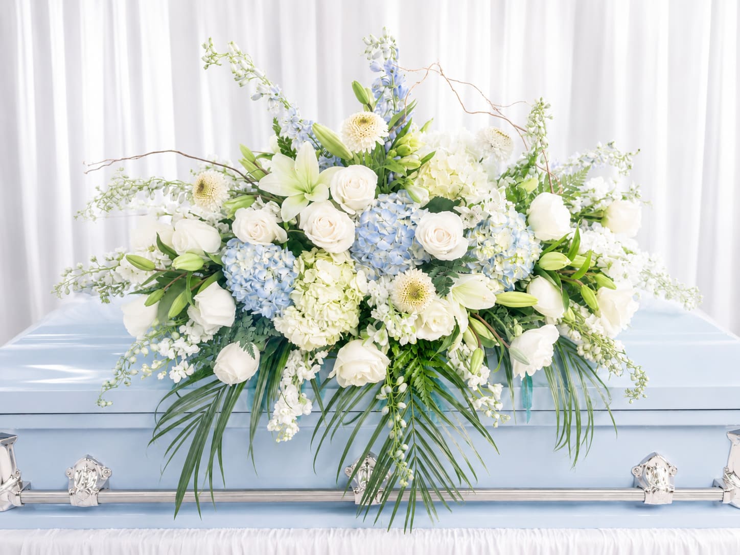 Heaven’s whisper - A serene, large low arrangement in soft blue and white that reads as both comforting and ceremonious - perfect as a wedding centerpiece or a sympathy arrangement. Cream roses and pale lilies take center stage to create an elegant, calm presence the person you're thinking of will find reassuring; the cool blue hydrangeas and small white accents add a gentle, airy contrast that keeps the design feeling fresh rather than heavy.  This expansive, layered composition is finished with arching branches and trailing greenery for a sweeping, formal silhouette. The piece is lush and structured, composed of roses, lilies, hydrangeas, chrysanthemums, daisies and other small blooms in a harmonious white, blue and green palette. Sized to make a statement on a large table or altar, it offers visual depth through varied textures - silky petals, rounded hydrangea clusters, and delicate filler blooms - so it reads beautifully from every angle.  For a wedding, it brings refined elegance to head tables or ceremony settings; for sympathy, its calm tones and generous scale convey heartfelt sympathy and steady support. Care is simple: keep the arrangement in a cool spot away from direct sun and replenish water as needed to maintain freshness through the important moments it will mark.   If any design elements are of major importance to your order, please include them in the florist instructions at checkout or contact us to ensure availability.