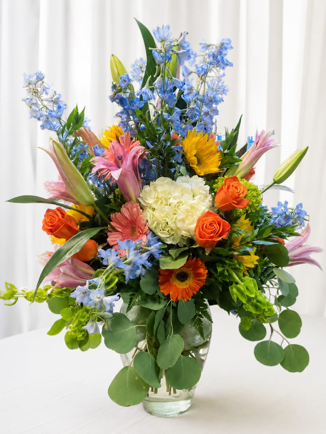 Sam’s blooms - A bright, garden-like bouquet that feels like a celebration in bloom-this large flower arrangement features lilies, roses, gerbera daisies, hydrangeas and delphinium arranged for maximum color and presence. Perfect as a birthday bouquet, a congratulations gift, or a general celebratory floral surprise, Sam's blooms is designed to make the person you're thinking of feel noticed and genuinely celebrated.  Tall blue delphinium rises above pink lilies and pink gerbera, while vibrant orange roses and a pale white hydrangea cluster add contrast and texture in the center. The mixed bouquet arrives in a clear glass vase, creating an upright, abundant silhouette that works well on a dining table, office desk, or entryway. The lively mix of blue, pink, orange and white gives a cheerful, uplifting look that reads joyful from across the room.  Choose this arrangement when you want someone to feel brightened and honored-ideal for milestone birthdays, promotions, or simply saying "I'm proud of you." As a large design, it fills a room; order details and delivery options are available at checkout to ensure it arrives fresh and ready to impress.   If any design elements are of major importance to your order, please include them in the florist instructions at checkout or contact us to ensure availability.
