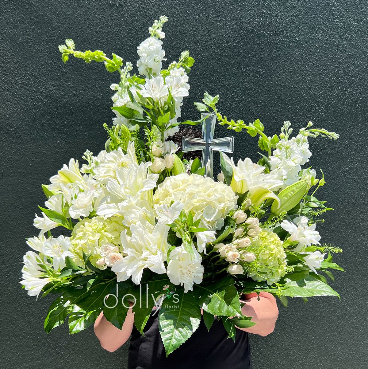 Peaceful Cross - A luminous arrangement built around pure white lilies and airy hydrangeas creates a serene focal point for a funeral or memorial service. This large sympathy arrangement features a clear glass cross pick rising from the center, making it an unmistakable tribute that expresses reverence and gentle remembrance.  Layered with white carnations, alstroemeria and small roses, the rounded composition is full and balanced, with bright green accents and glossy foliage that give the piece fresh contrast and texture. The formal, white-dominant palette reads as calm and respectful while the mix of blooms adds subtle scent and soft movement suitable for a ceremony or graveside tribute.  Intended to comfort the person you're honoring, this design conveys sympathy and deep respect-an appropriate choice for a service, visitation, or memorial table. As a large piece, it arrives ready to display; keep it in a cool location away from direct sun and replenish water as needed to extend freshness.