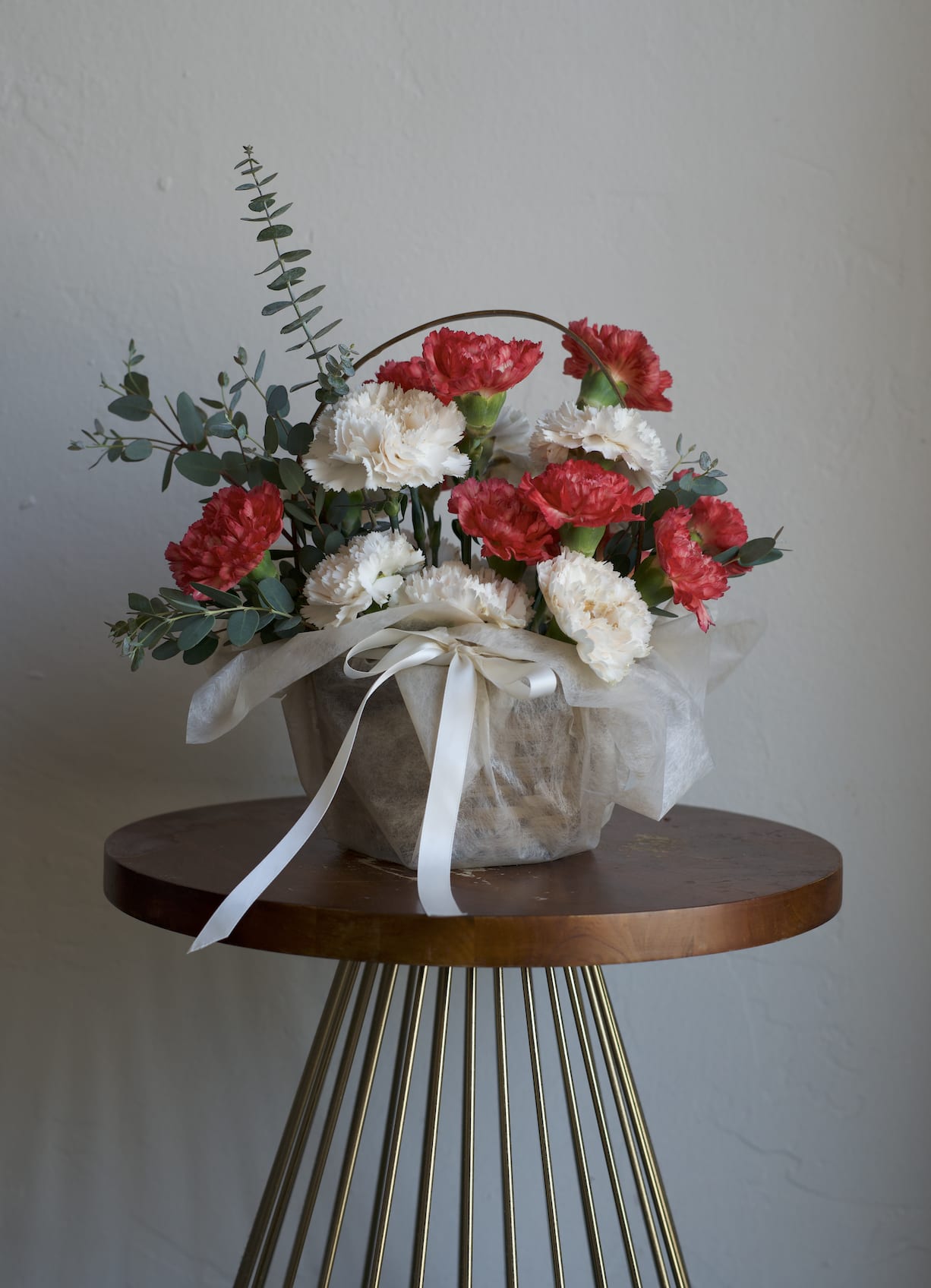Basket of Blooms - A cheerful cluster of red and white carnations in a low woven basket creates an immediate, heartfelt impact-the perfect flower arrangement for a birthday bouquet, anniversary roses alternative, or a simple thank-you gift.  This Basket of Blooms pairs ruffled carnation heads with airy eucalyptus sprigs for cool green contrast and light height, keeping the composition informal but polished. The arrangement sits low in the basket, wrapped in soft white tulle and tied with a white ribbon for a charming, hand-delivered look that's easy to display on a table or desk.  Ideal for celebrating small victories, saying "I'm thinking of you," or brightening someone's day, this keepsake-style basket feels thoughtful without being over the top. 