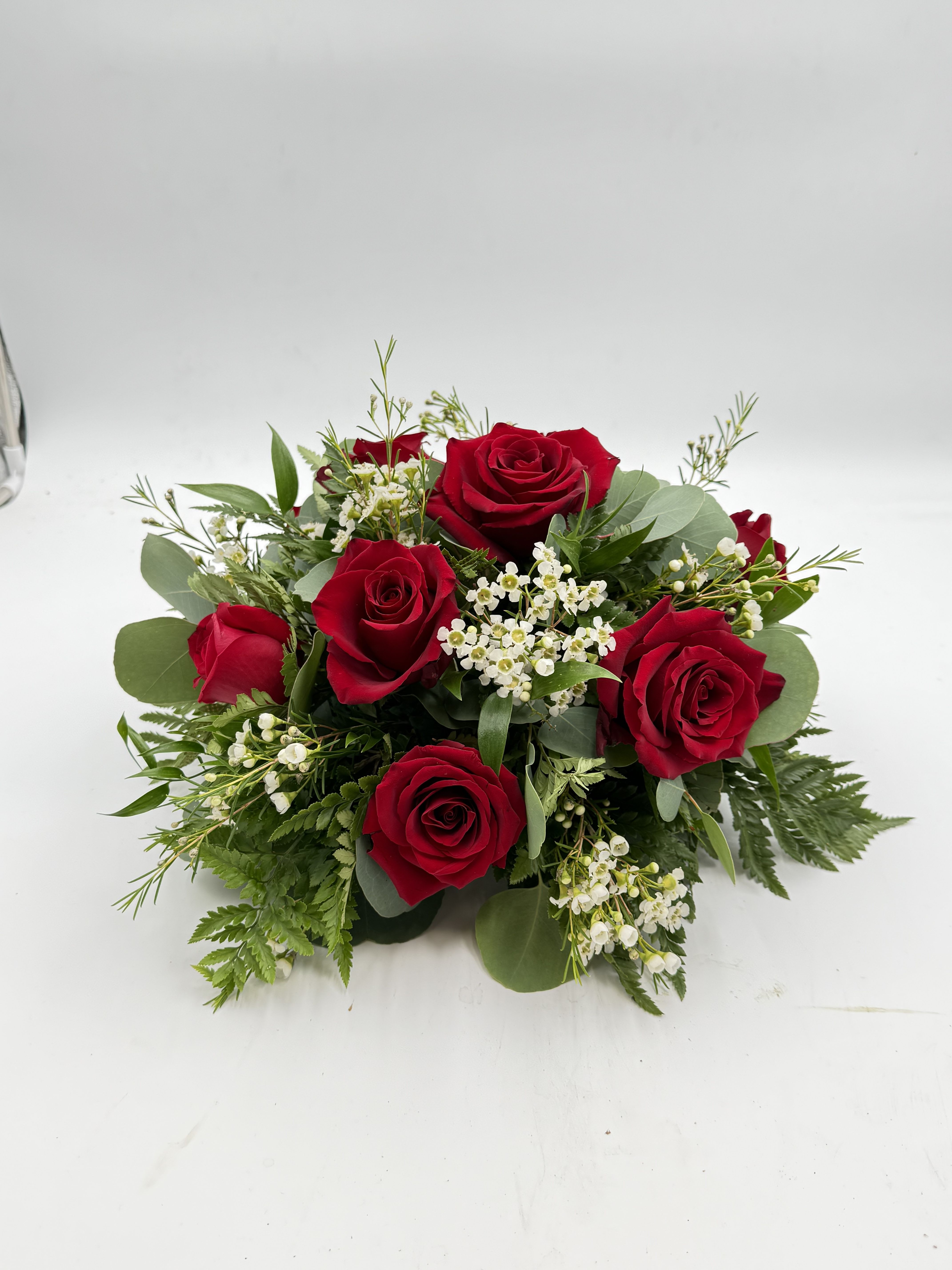 Classic Rose Centerpiece - Deep clusters of deep red roses form the heart of this low, rounded flower arrangement-perfect as a romantic anniversary roses centerpiece or a formal wedding table arrangement. The rich red-and-white palette reads classic and elegant, so the person you're thinking of will feel truly celebrated whether this is set at a dinner table, used as a sympathy tribute, or placed as a holiday focal point.  Small white filler blossoms are woven between the roses and framed by a mix of glossy and matte greens and fern-like foliage, giving the design a full, layered texture without rising tall above the table. The compact, medium hand-tied shape keeps blooms close to the surface so guests can converse across it; it looks polished on a white tablecloth alongside wine glasses, dinner plates, and neatly folded napkins.  Ideal for weddings, anniversaries, formal dinners, or a quiet yet meaningful gift, this centerpiece sends a message of timeless affection and refined taste. The balanced, neat presentation needs only basic care: keep stems in fresh water, trim occasionally, and swap water every few days to maintain that lush, just-arranged look through the celebration.