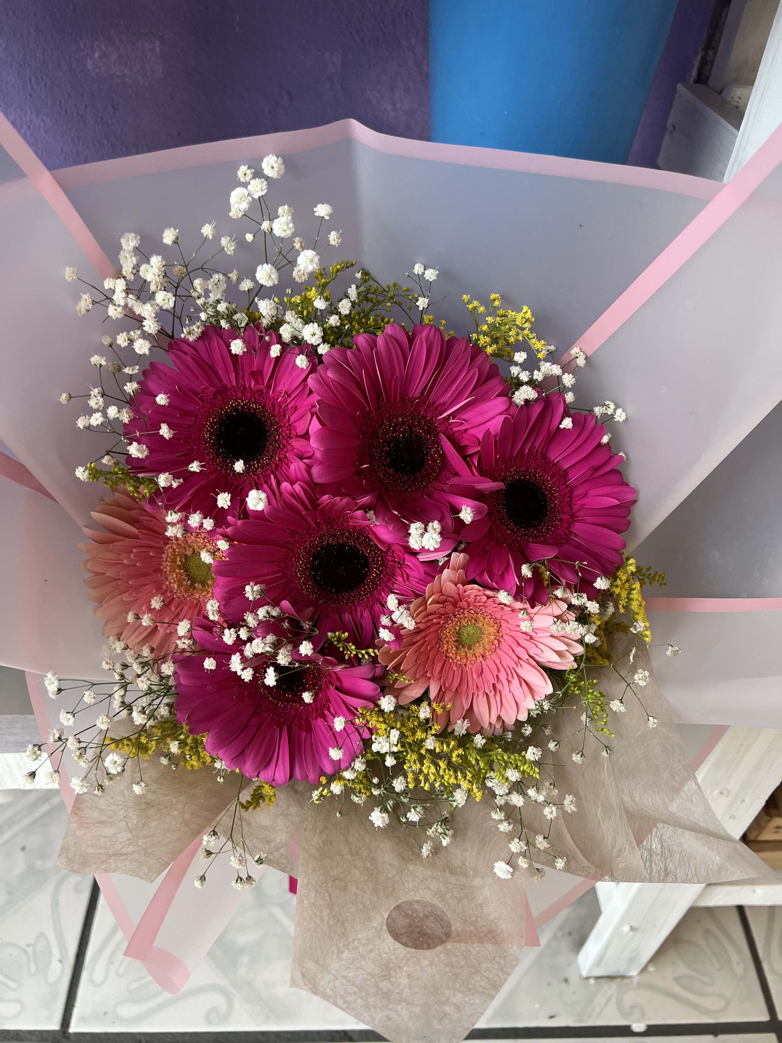 Gerbera Bouquet - Bold, bright, and impossible to ignore — this stunning bouquet features lush magenta and soft pink gerbera daisies, each bloom wide and full, surrounded by delicate baby's breath and cheerful yellow solidago. Wrapped in a beautiful layered blush and kraft paper with a pink satin ribbon, it's the ultimate mood-lifting gift. Whether celebrating a birthday, graduation, promotion, or just spreading some joy, this bouquet delivers maximum color and happiness in every stem.