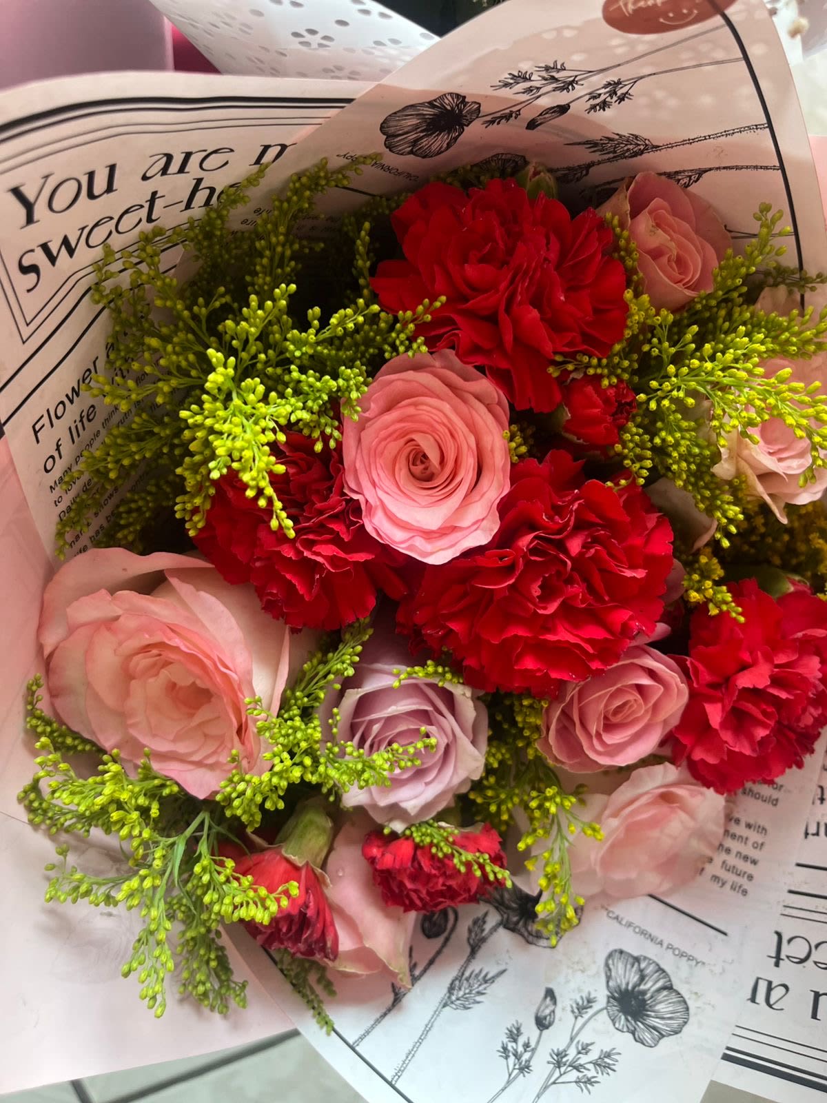 Sweetheart Bouquet — Pink &amp; Red Mix - A lush, romantic hand-wrapped bouquet bursting with soft pink roses in two tones, bold red carnations, and bright yellow-green solidago — all beautifully presented in a decorative botanical-print wrap. The contrast of delicate blush and warm crimson creates a bouquet that feels both playful and deeply heartfelt. Whether you're celebrating a birthday, anniversary, or just saying "I'm thinking of you," this arrangement delivers color, texture, and charm in every stem. Perfect for gifting on the go or as a stunning centerpiece.
