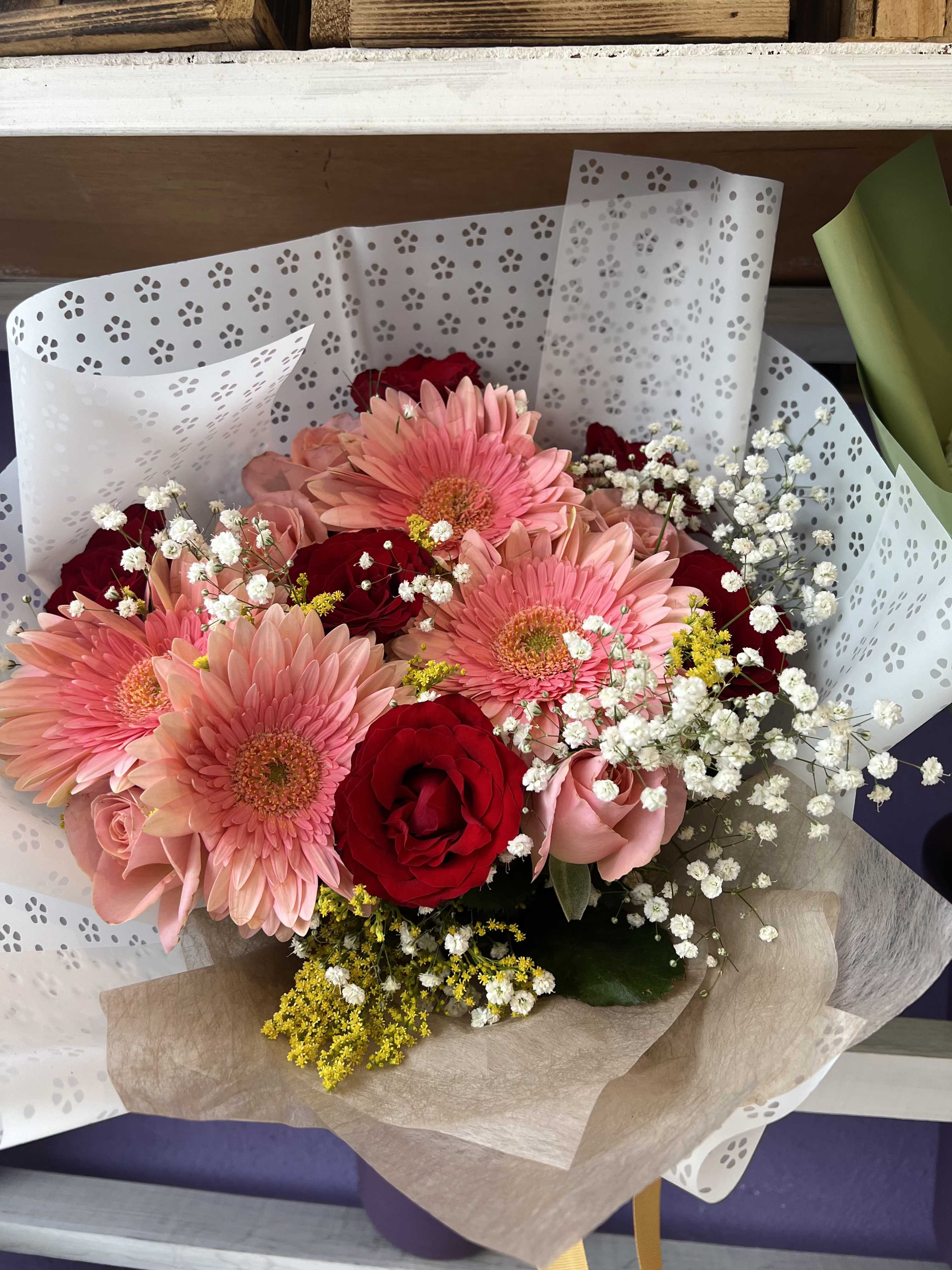 Blushing Garden Lace Bouquet - Sweet and utterly charming — this full, lush bouquet pairs soft salmon-pink gerbera daisies with deep red and blush roses, accented by delicate baby's breath and warm yellow solidago. The real showstopper is the wrapping: a beautiful laser-cut lace paper that gives this bouquet a romantic, heirloom quality that feels truly special from the moment it's received. A perfect balance of playful and romantic, this is the go-to gift for birthdays, Mother's Day, anniversaries, bridal showers, or anytime you want to make someone feel genuinely celebrated.