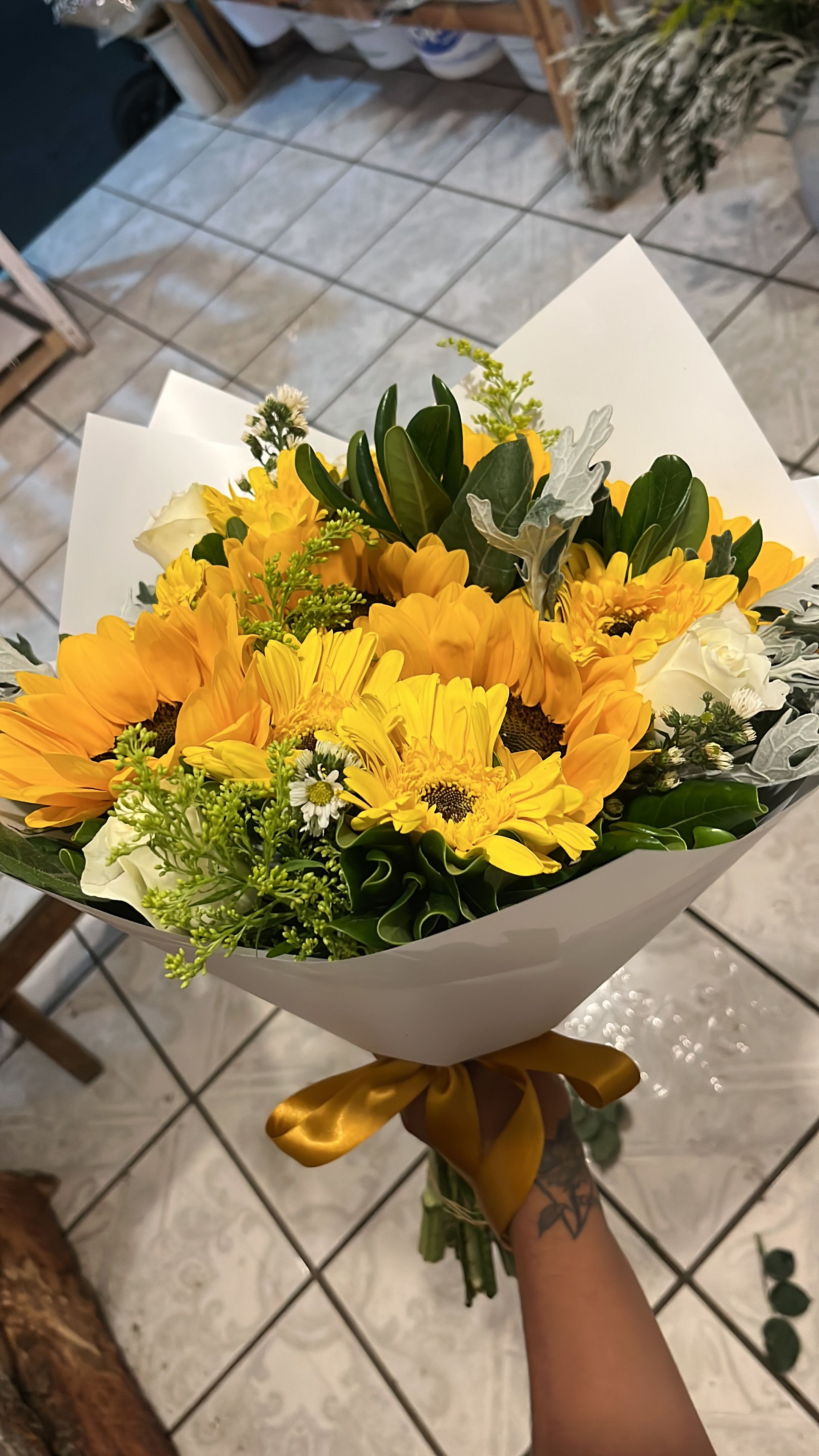 Sunshine Garden - Pure joy wrapped in white — this radiant bouquet combines oversized golden sunflowers, cheerful yellow gerbera daisies, and soft white roses with lush tropical foliage, silvery dusty miller, yellow solidago, and delicate white daisy mums, all tied with a rich gold satin ribbon. The layered mix of textures and bloom sizes creates a bouquet that feels abundant, garden-fresh, and utterly alive. The ultimate expression of warmth, friendship, and celebration — perfect for birthdays, teacher appreciation, get well, graduations, or brightening any space that needs a little more sunshine. A smile in bouquet form.