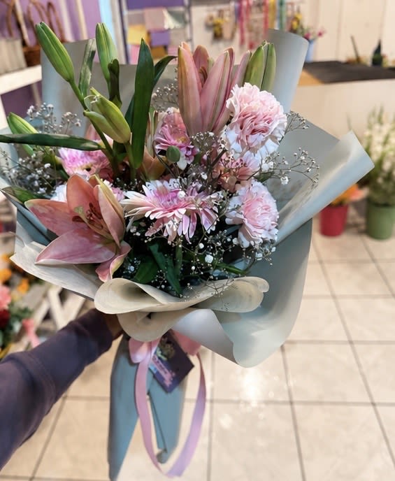 Pink Lily Elegance Bouquet - Graceful and fragrant, this beautifully structured bouquet features tall pink Oriental lily stems — several buds ready to unfurl over the coming days — alongside soft pink carnations, a blush gerbera daisy, and delicate baby's breath with silver accents. Wrapped in a sophisticated dusty blue layered paper with a soft pink satin ribbon, the cool wrap makes the warm pink blooms absolutely glow. A bouquet that keeps blooming long after it's received — perfect for Mother's Day, get well wishes, sympathy, or simply to show someone you're thinking of them.