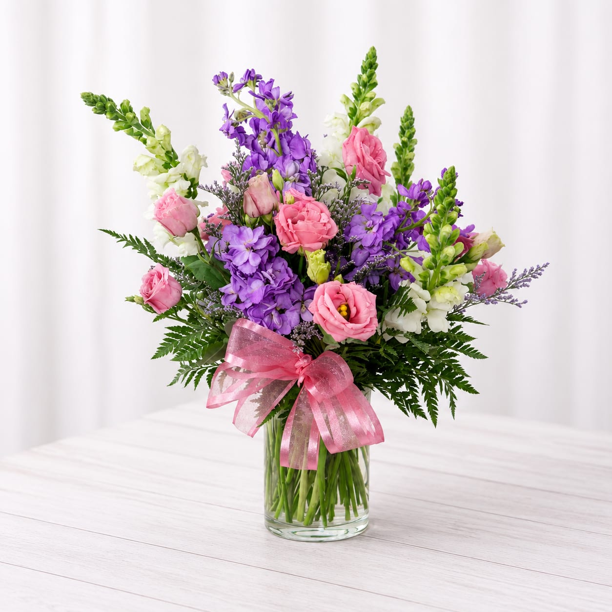 Sweetheart Statement - Tall white snapdragon spikes crown this Sweetheart Statement flower arrangement, giving it a dramatic vertical silhouette that feels both romantic and celebratory. As a birthday bouquet or anniversary arrangement meant for someone special, it reads as heartfelt and joyful-the pink ribbon-tied clear glass vase keeps the look fresh and approachable while the color palette of pink and  purple sends a classic message of love and warmth.