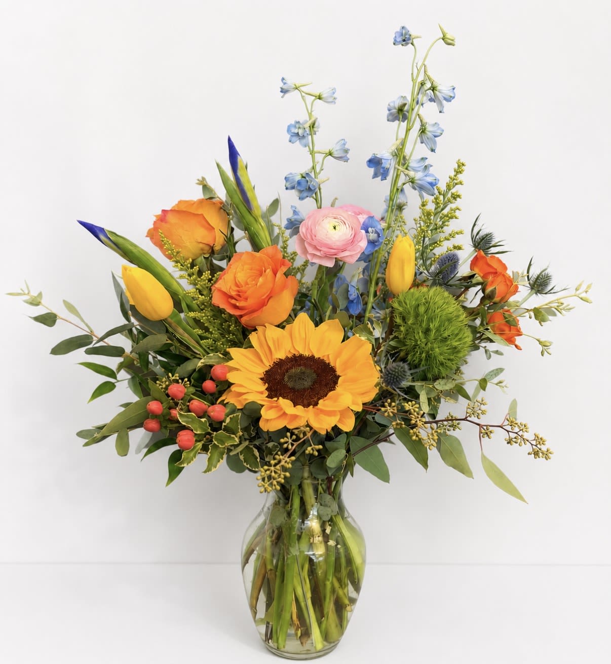 Kansas Cottage - A bold sunflower takes center stage in this mixed-color flower arrangement, making the Kansas Cottage flower arrangement perfect for a cheerful birthday bouquet, a warm Mother's Day surprise, or a just-because pick-me-up. Bright oranges and sunny yellows meet soft pinks, whites, and purples to create a celebratory look that also suits congratulations, graduation, or a thank-you gift-and its energetic palette works year-round from spring and summer through cozy autumn occasions like Thanksgiving and Labor Day.  This garden-style design pairs large sunflowers with lilies, iris, roses and spray roses, snapdragons, tulips and smaller yellow accent blooms, arranged in a clear glass vase that shows the stems and keeps the feel fresh and casual. Tall spires and outward-reaching stems give it a lively, asymmetrical silhouette while airy greenery and bright filler add texture and movement. The result is a vibrant, feel-good bouquet that will make the person you're thinking of feel genuinely celebrated; vase included and ready to display-no extra fuss, just unwrap and brighten their day.