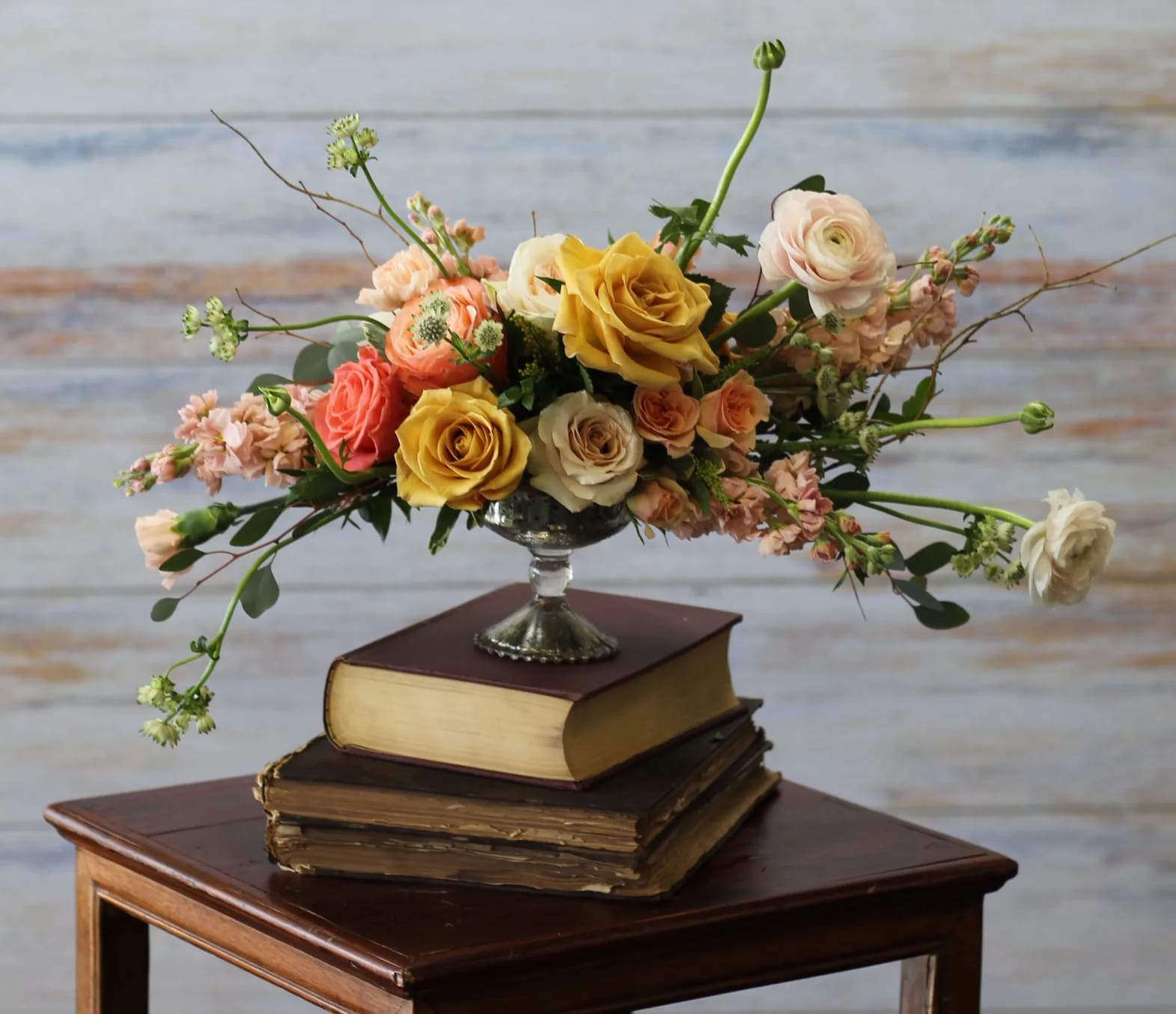 Low arrangement of yellow and peach roses and ranunculus in a silver pedestal bowl on stacked books.