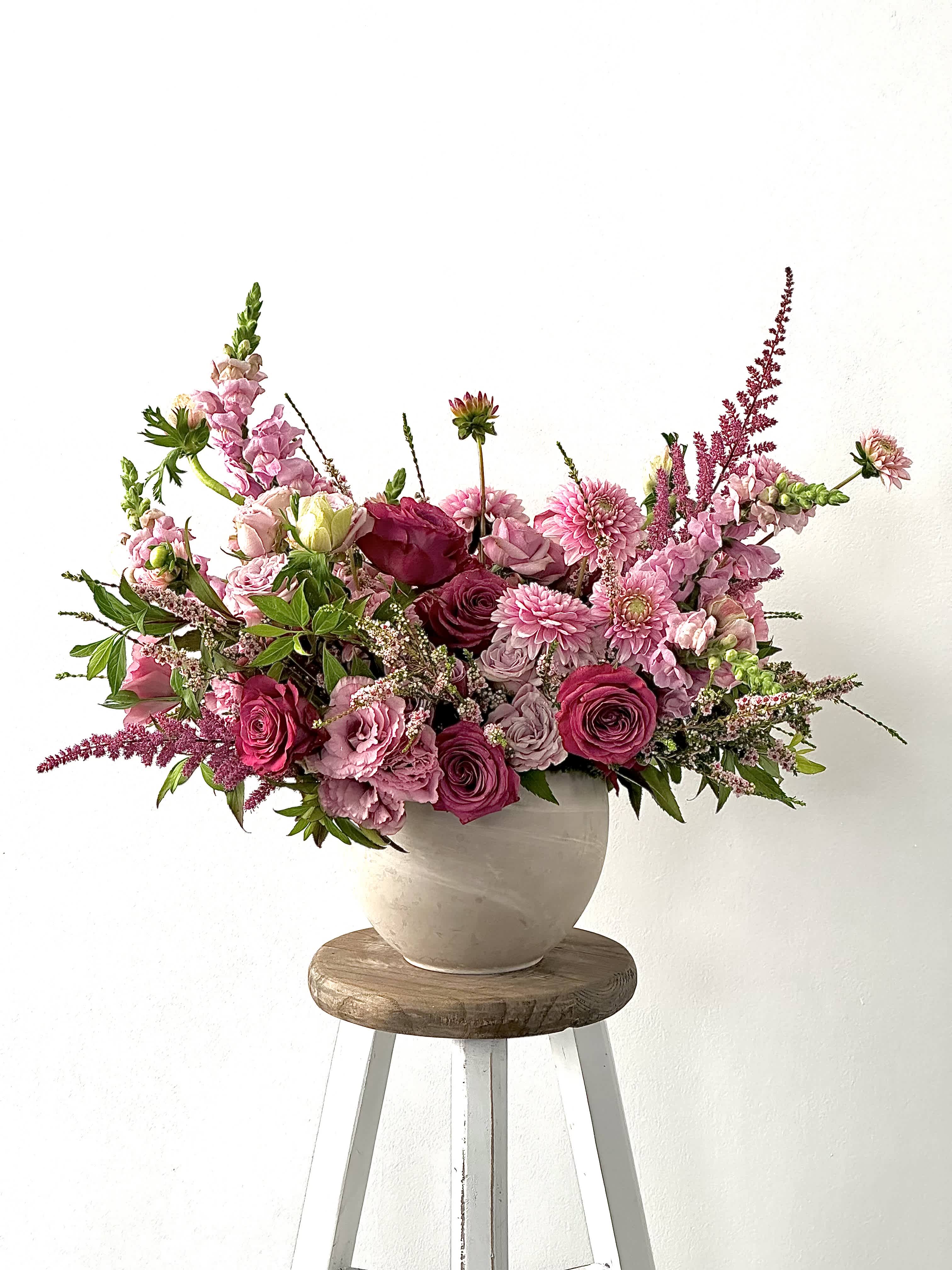 Pink mixed flower arrangement in a beige vase on a wooden stool