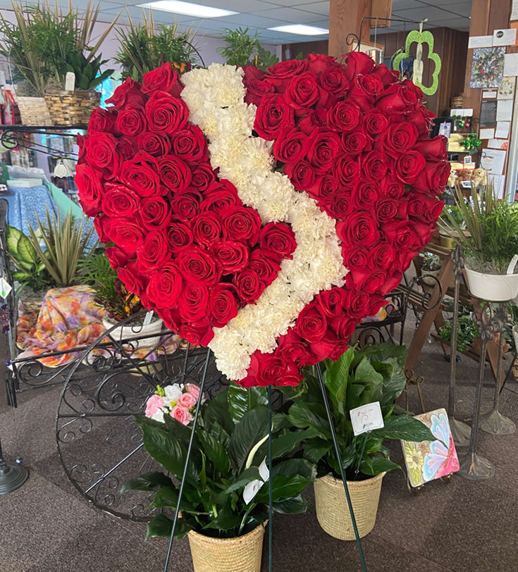 Tall heart-shaped standing spray of red roses with a white floral stripe above two potted green plants