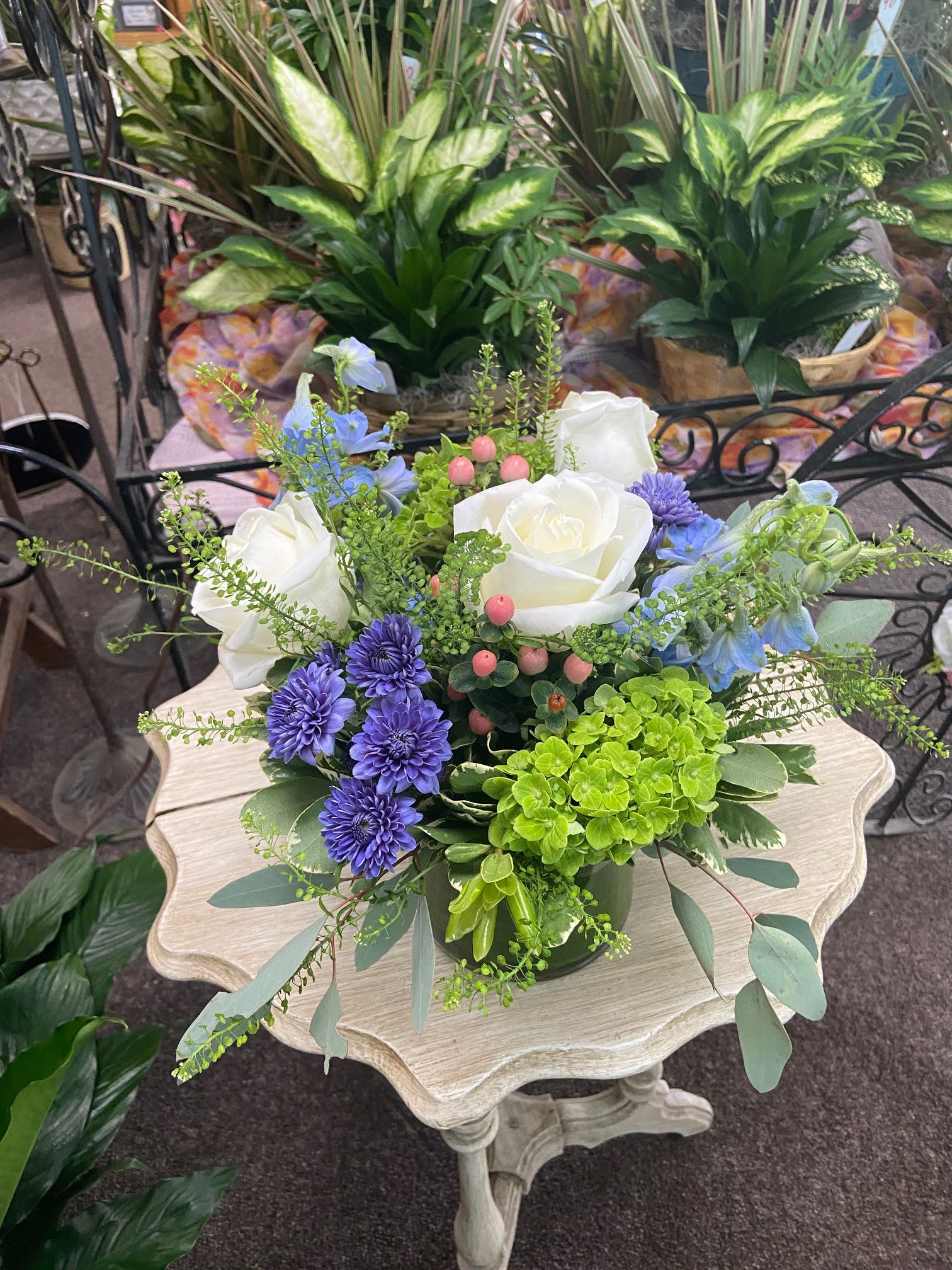 Low arrangement of white roses, purple mums, blue blooms, and green hydrangea in a vase on a small table