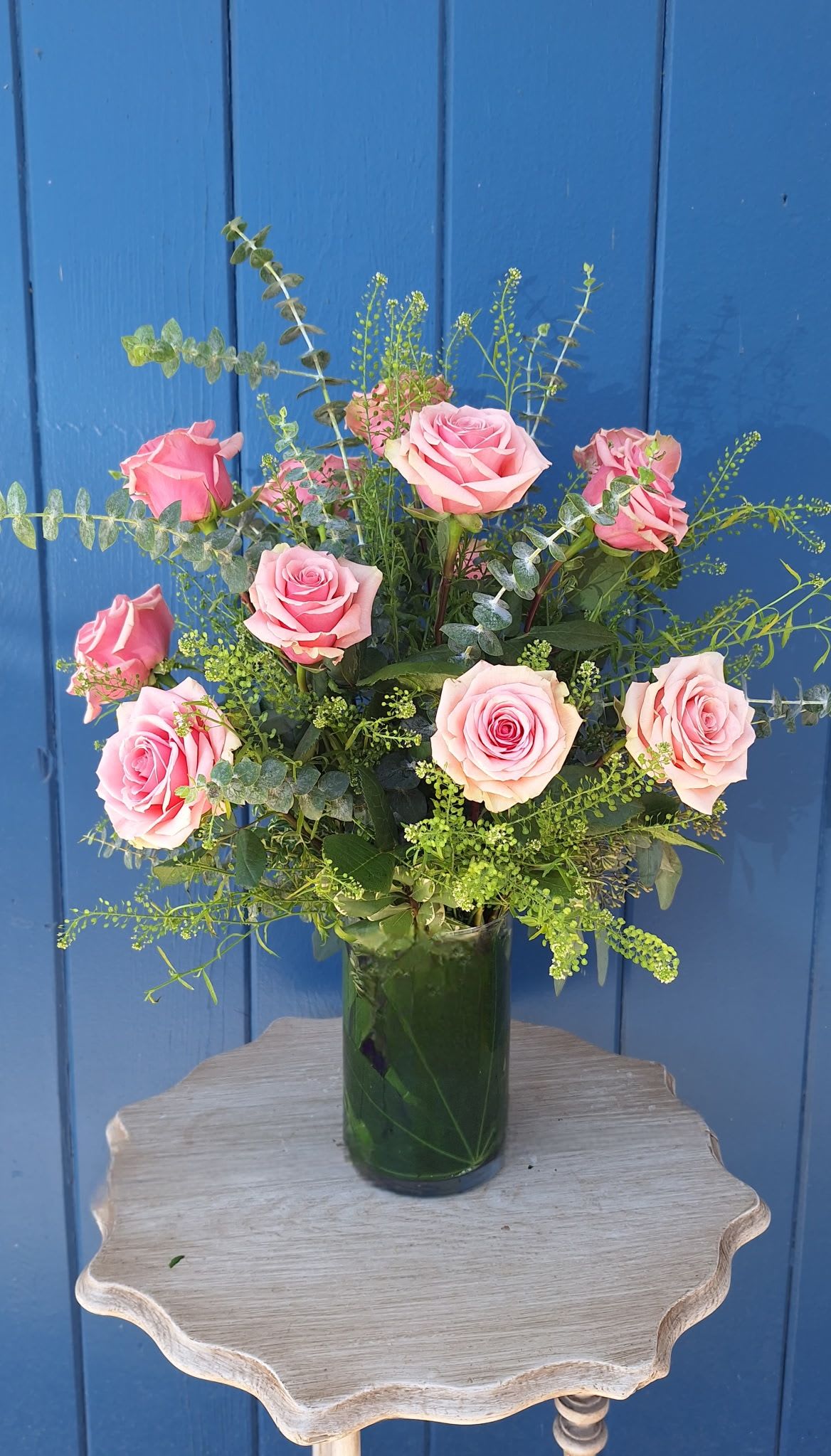 Tall arrangement of pink roses with airy filler in a clear glass vase on a small wooden table