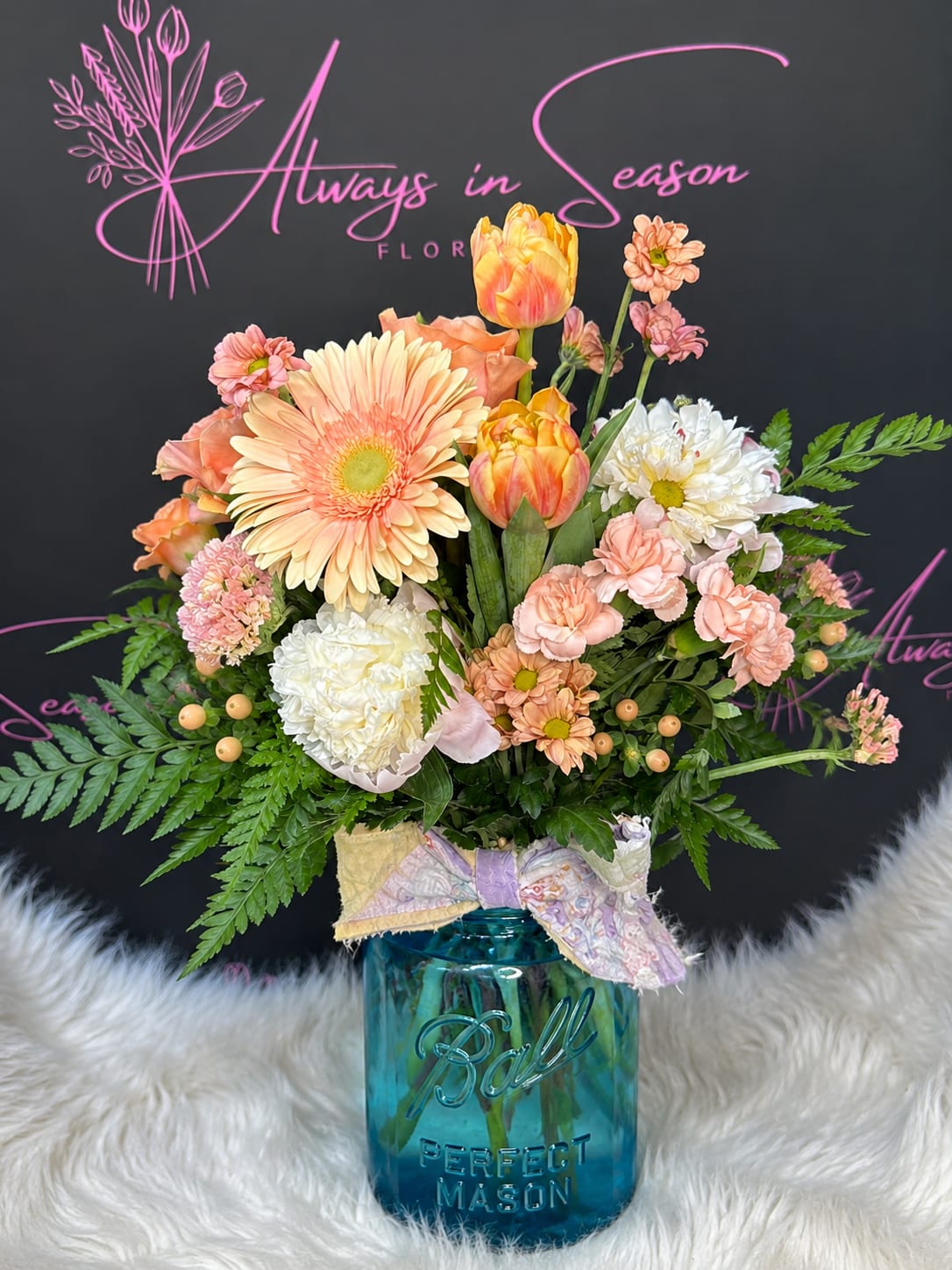 Heirloom Peach Garden - Peach and blush gerbera daisies and tulips take center stage in this spring-inspired flower arrangement, creating a soft, cheerful bouquet perfect as a birthday bouquet, thank-you gift, or just-because surprise. The arrangement reads fresh and feminine-an easy way to make the person you're thinking of feel warm, noticed, and celebrated.  Clusters of peach carnations and white daisy-like chrysanthemums fill out the rounded, garden-style design, while dense green foliage adds texture and a lush backdrop. The stems are arranged in a blue glass mason jar and finished with a tied fabric ribbon for a casual, handcrafted look that suits kitchen counters, office desks, or bedside tables.  This arrangement is designed to feel full and abundant without being fussy-ideal for spring celebrations or everyday display. Care is simple: keep the water fresh and trim stems periodically so the blooms stay lively; the mason jar vase makes it easy to place and replenish.