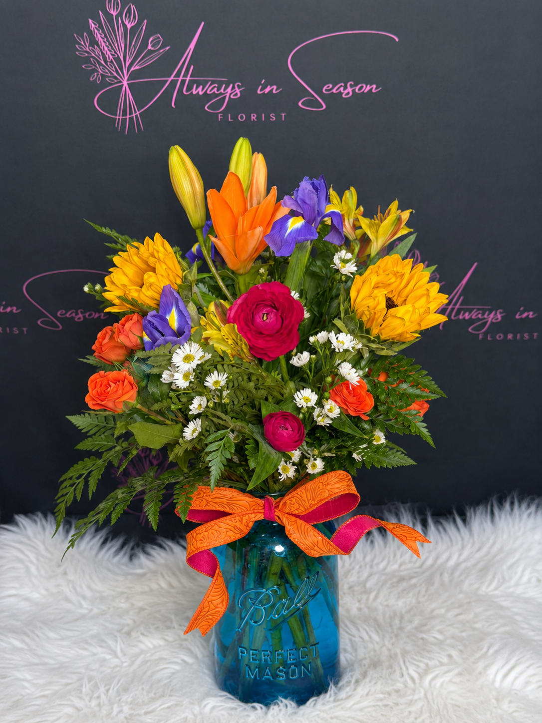 Grandma’s Vintage Garden Jar - Bursting with sunshine and vintage charm, this flower arrangement is a joyful everyday gift that says "thinking of you" in the most colorful way. Perfect as a birthday bouquet, a congratulations surprise, or a cheerful thank-you for a loved one, the mix of lilies, irises, roses, daisies, and ranunculus creates an exuberant, hand-tied look that feels both festive and familiar-like a memory of Grandma's garden brought to the table.  Bright orange lilies and orange roses meet vivid pink ranunculus and hot pink roses, while purple irises and sunny yellow daisy-like blooms add lively contrast; touches of small white filler flowers give the composition a delicate, vintage texture. The flowers are densely arranged in a full, rounded design and presented in a blue mason jar finished with a wide orange ribbon for a casual, handcrafted look that fits cottage, farmhouse, or eclectic décor.  Made to make the recipient feel truly celebrated, this arrangement reads as upbeat and energetic-ideal for brightening someone's day or marking a happy milestone. The compact, upright design is easy to place on a kitchen table or desk; swap the water every few days, recut stems at an angle, and keep away from direct heat to help the blooms last longer.