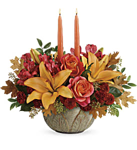 Artistic Glow Centerpiece - Make the season glow with this bright and bountiful fall bouquet of lilies and roses, arranged in a hand-glazed, oven-to-table stoneware bowl--the perfect mix of pretty and practical! This glowing arrangement includes orange roses,  asiatic lilies, red alstroemeria, bronze button spray chrysanthemums, burgundy cushion spray chrysanthemums, seeded eucalyptus, huckleberry, yellow transparent oak leaves, and two peach candles. Delivered in an Artisanal Autumn Bowl. Approximately 17 1/2" W x 15 1/2" H