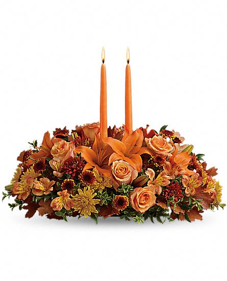 Thanksgiving Centerpiece - This bright autumn centerpiece spins the magic of the Fall season with each sun-kissed petal to set your gathering space aglow with blooming beauty. Two taper candles are surrounded by a gorgeous arrangement consisting of  Asiatic lilies, gerbera daisies and roses with golden Peruvian lilies, burgundy cushion poms and assorted greens to create the perfect addition to your Autumn celebration. BETTER centerpiece is approximately 8"H x 16"D. Approx. 8H x 15W