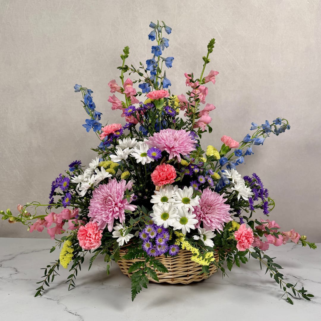 Pastel Peace Basket - Soft, garden-fresh colors and a gentle, arching silhouette make this flower arrangement a comforting way to say "I'm thinking of you" or to offer sympathy. The Pastel Prayers Basket arranges pink carnations and lavender cremones with clusters of white daisies at its heart, while tall blue delphinium and pink snapdragons rise above for graceful height. The overall look reads warm and uplifting, perfect for a tasteful gift for the spring.  Rendered in a light wicker basket, the mix includes purple monte casino and yellow statice filler flowers scattered throughout for pops of contrast and a meadow-like texture. The pastel color palette keeps the design both soothing and cheerful, and the fan-shaped composition offers an open, garden-inspired feel. Sized to make a visible, comforting statement without overwhelming a room, this arrangement arrives ready to display.  Dimensions approx. L 32" x H 26"