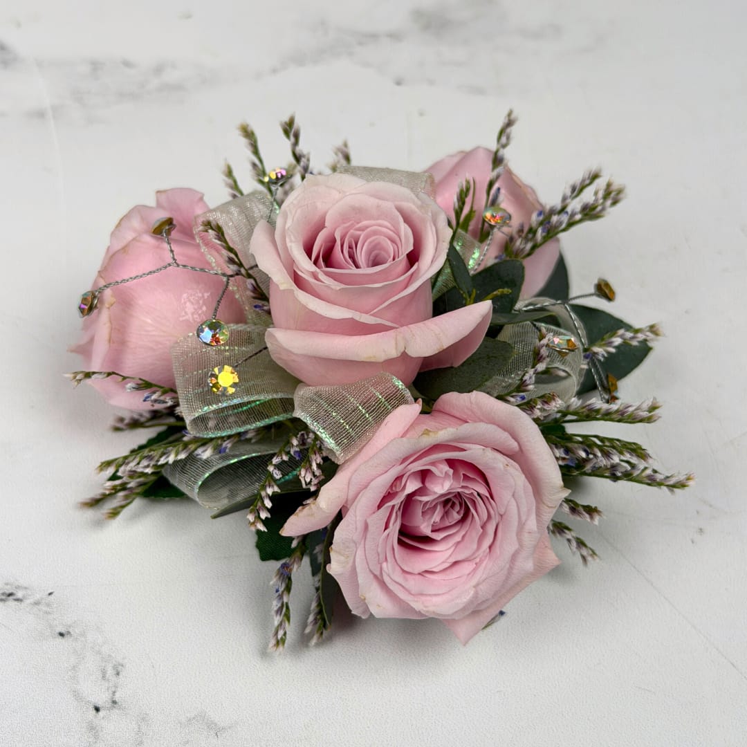 Amethyst 4 Rose Wrist Corsage - Four pale lavender roses form a delicate, wearable cluster in this Amethyst 4 Rose Corsage, perfect for prom. The soft lavender, white and green palette reads delicate and refined, the kind of piece that makes the person you're thinking of feel elegant and cherished on their big night.  The corsage pairs four compact roses with fine filler, finished with sheer iridescent ribbon loops and small iridescent rhinestone gems for a touch of sparkle. Hand-assembled to sit comfortably on the wrist with a white lace wristlet, it's small, neatly constructed, and designed to coordinate with formalwear and pastel palettes. Light misting and keeping it cool until wear helps maintain bloom freshness.