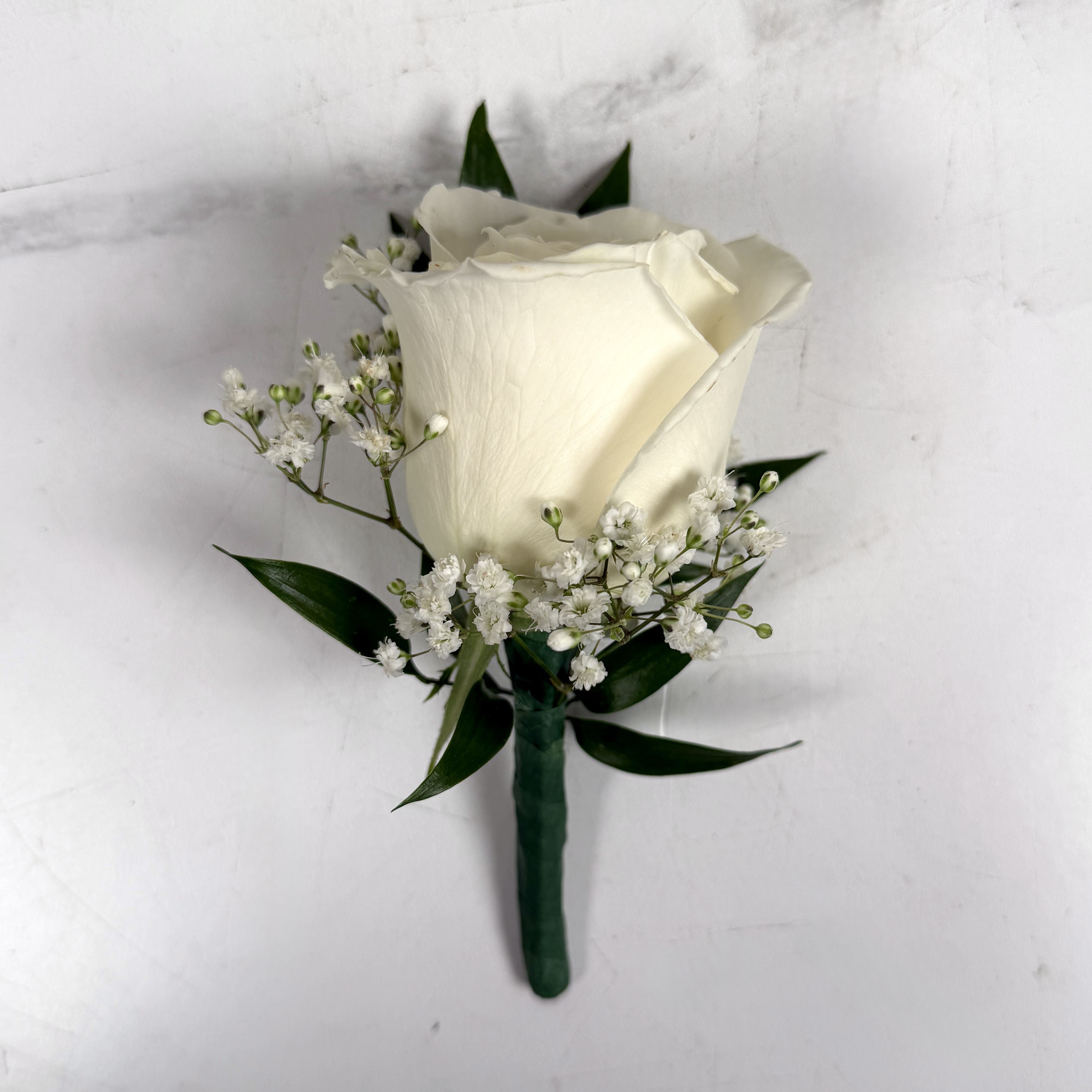 White Standard Rose Baby's Breath Boutonniere - One white standard, full-sized rose with baby's breath makes up this classic boutonniere, perfectly made to coordinate with virtually any outfit. The boutonniere is finished with a neat, floral taped boutonniere wrap around the stem and a boutonniere pin to attach the boutonniere for a clean, structured silhouette that sits comfortably on a jacket or suit.  To jazz up this boutonniere even more, consider adding on a special ribbon wrap or even some sparkle accents so you can really dazzle on the dancefloor. To select your colors, check out our add-ons below or at checkout and pick whichever color you want.