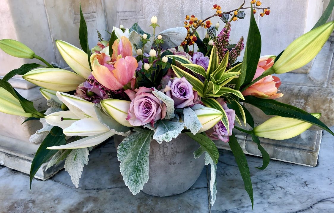 Low centerpiece of pink tulips, lavender roses, and white lilies in a gray pot