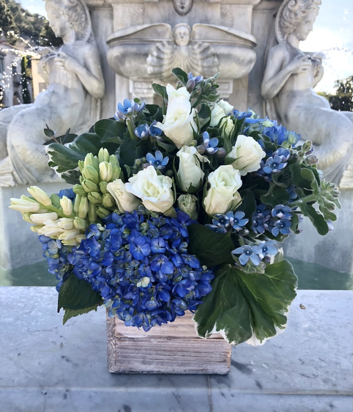 Compact blue and white flower arrangement with hydrangeas and roses in a rustic wooden cube vase.