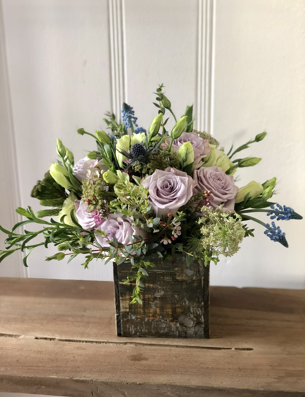 Lavender roses and white buds arranged with blue accents in a rustic wooden cube vase