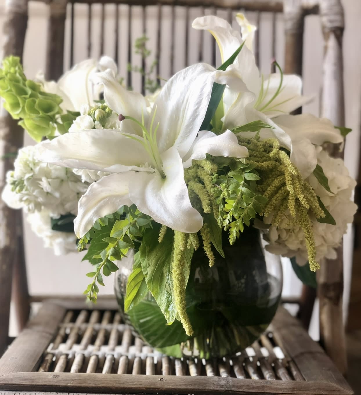 White lilies and mixed white and green flowers in a round glass vase.