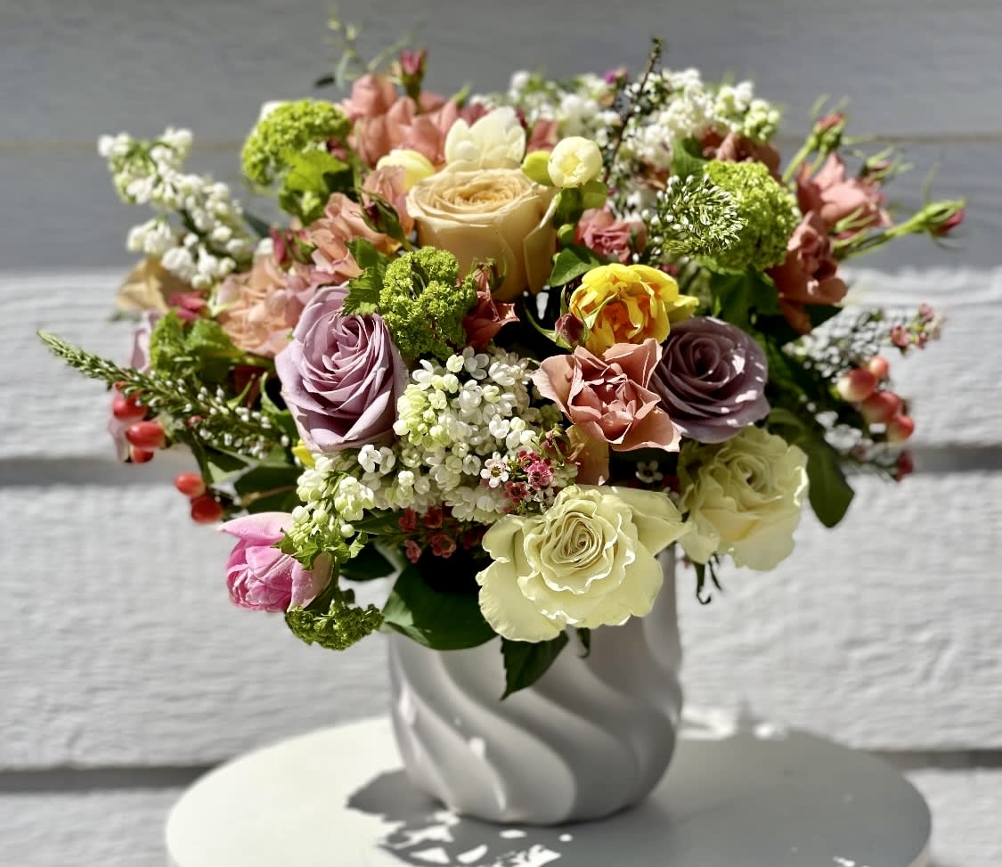 Mixed arrangement of cream, peach, lavender, and yellow roses with small blooms in a white ceramic vase