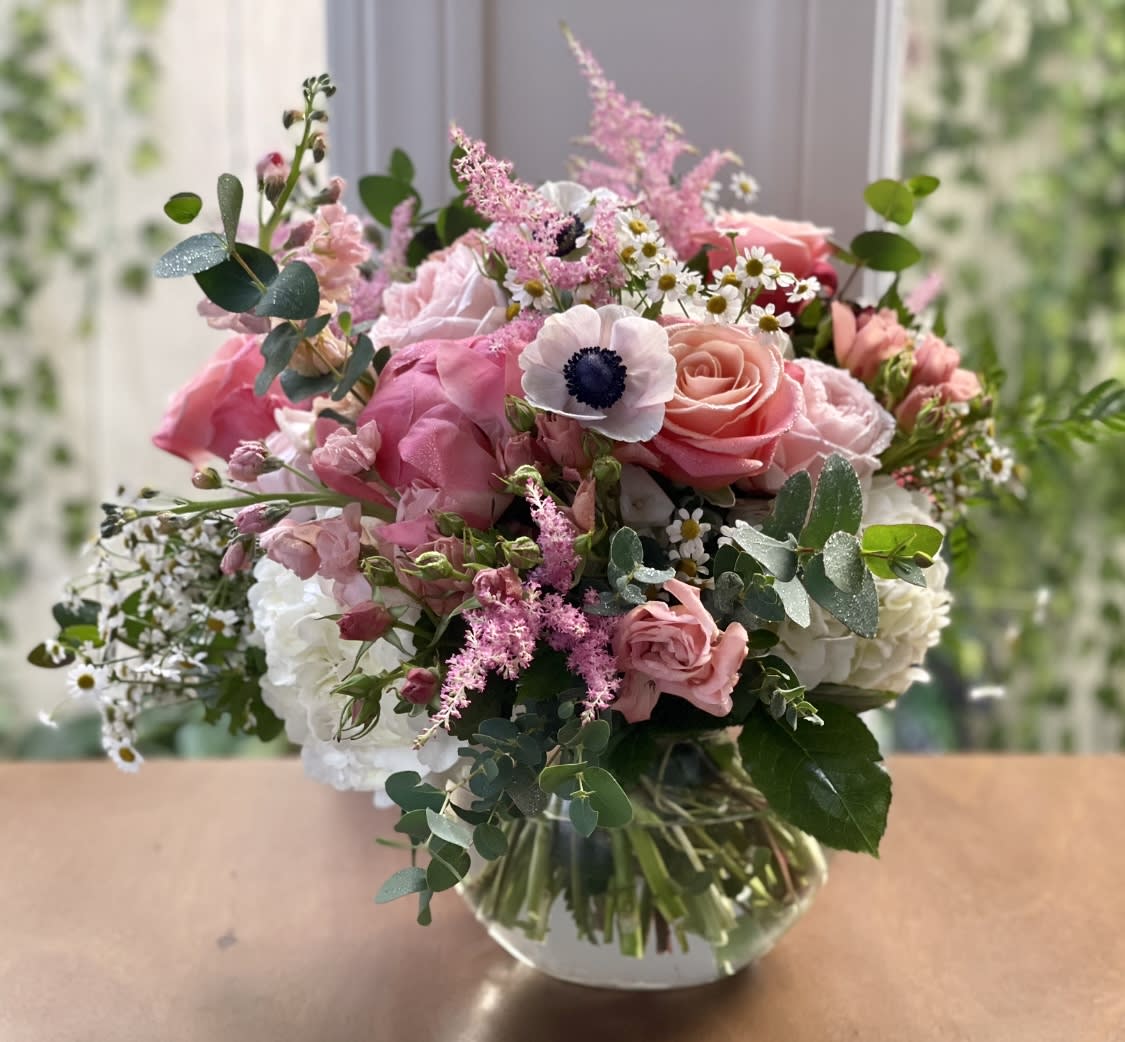 Pink and white mixed bouquet with roses and anemones in a round clear glass vase on a wooden surface.
