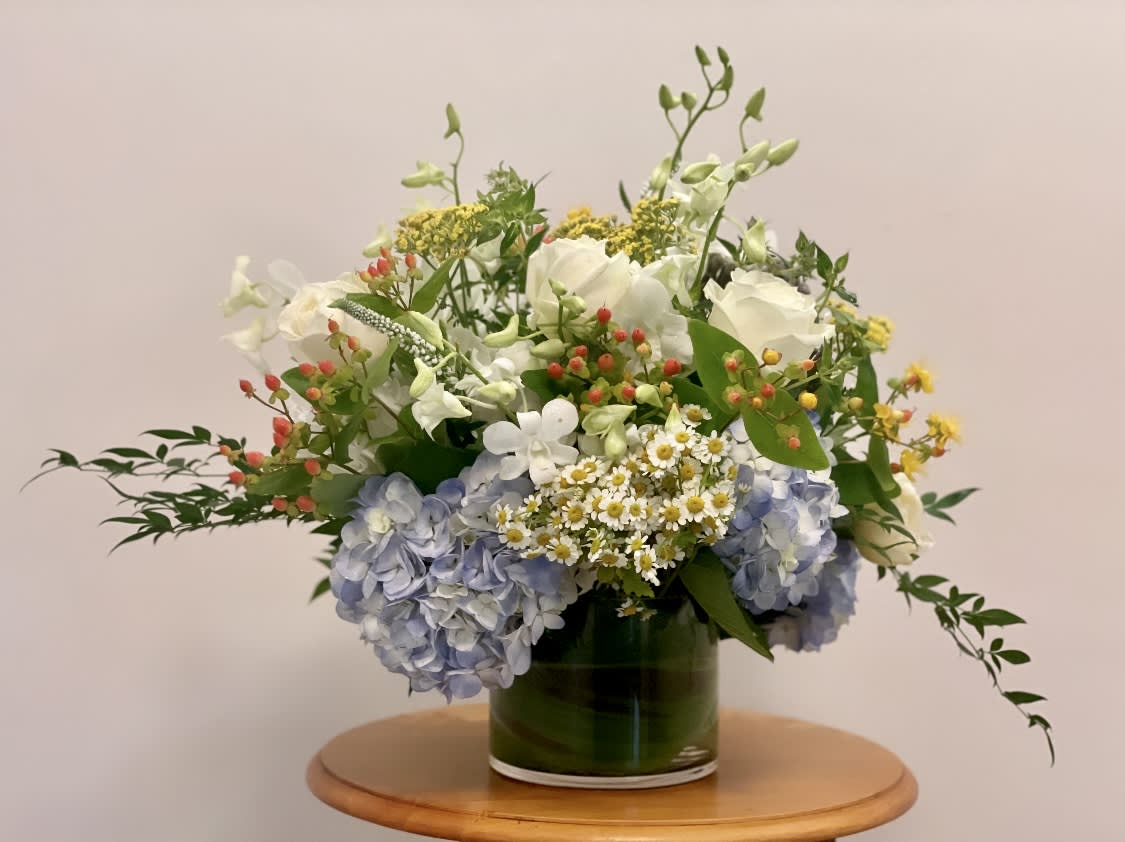 Low glass vase of white roses, blue hydrangeas, and small white daisies with berries on a wooden table