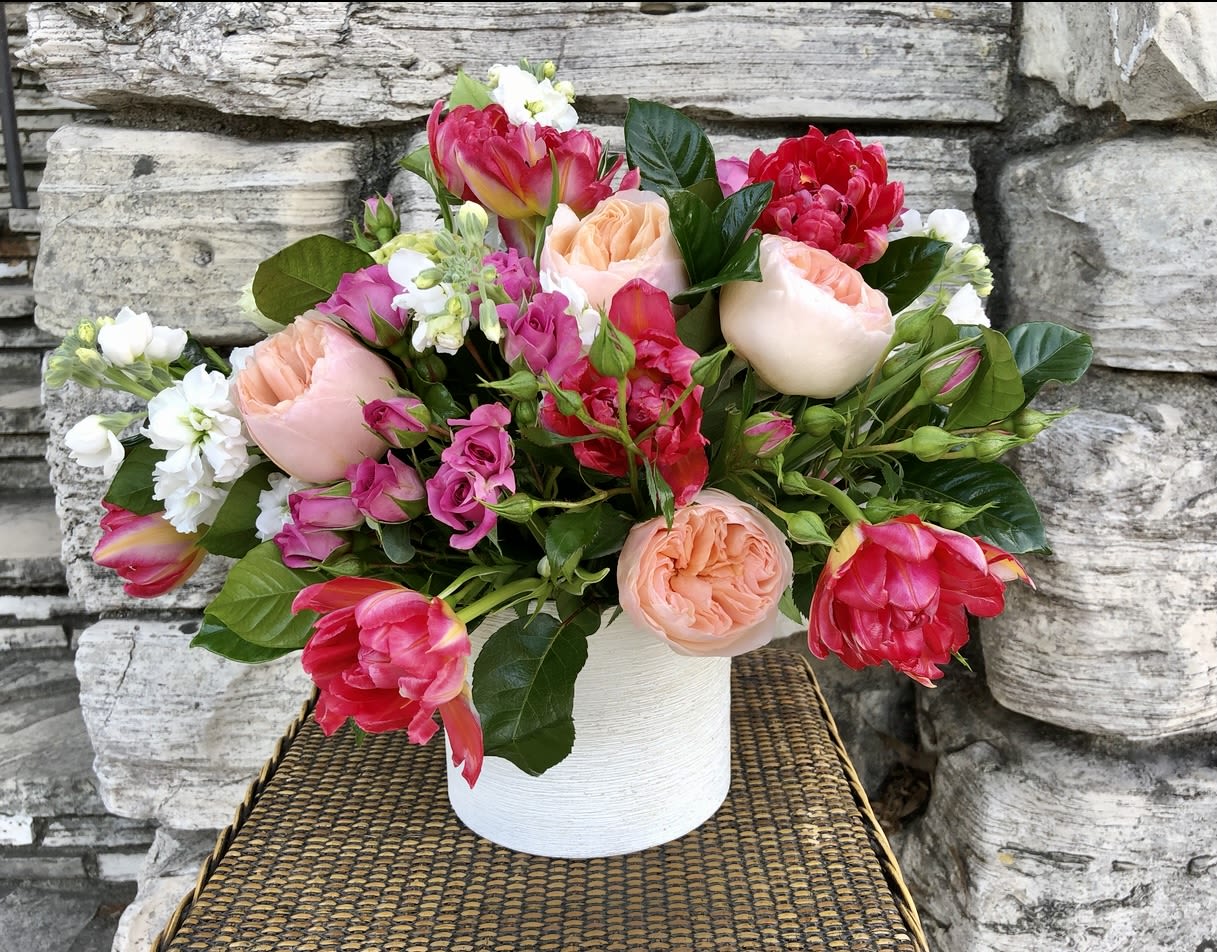 Low arrangement of pink and peach roses, tulips, and white stock in a white ceramic vase