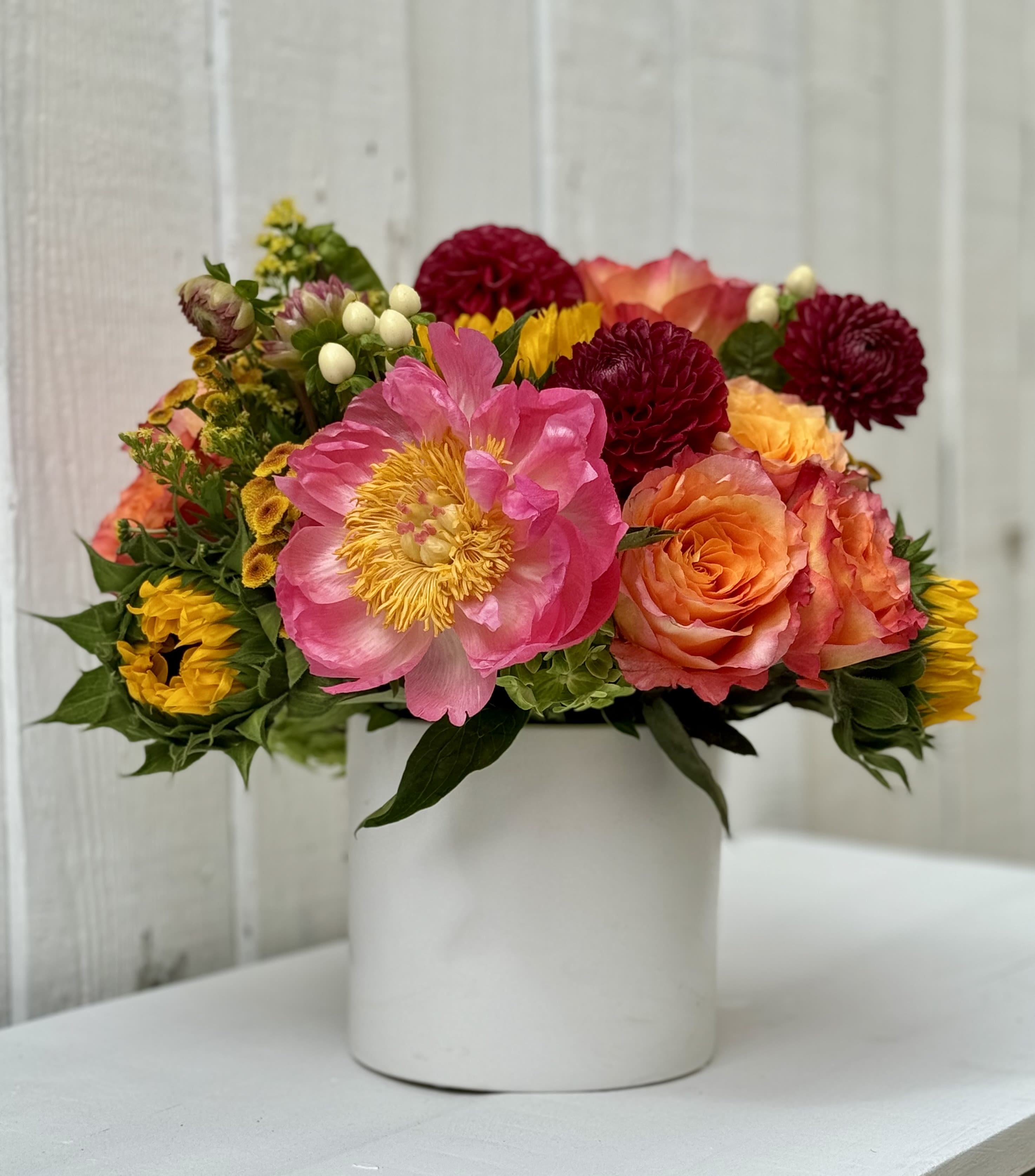 Bright arrangement of pink peony, orange roses, and yellow blooms in a white cylinder vase