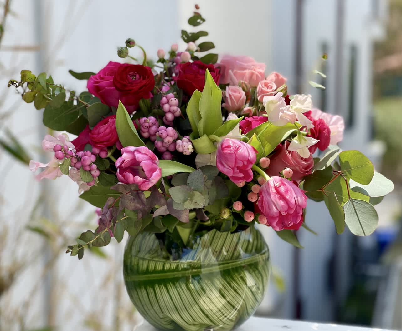 Bright pink and red tulips, roses, and ranunculus arranged in a round green glass vase