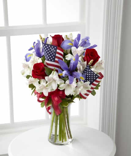 Unity Bouquet B30-4434 - Unity Bouquet sparks the hearts of all Americans with its patriotic beauty and dazzling color. Bright red roses mingle with blue iris arranged amongst white Peruvian lilies and assorted greens. Accented with two American Flags and a red, white and blue ribbon, this stunning bouquet arrives arranged in a clear glass vase to create a gorgeous way to celebrate our Independence Day.