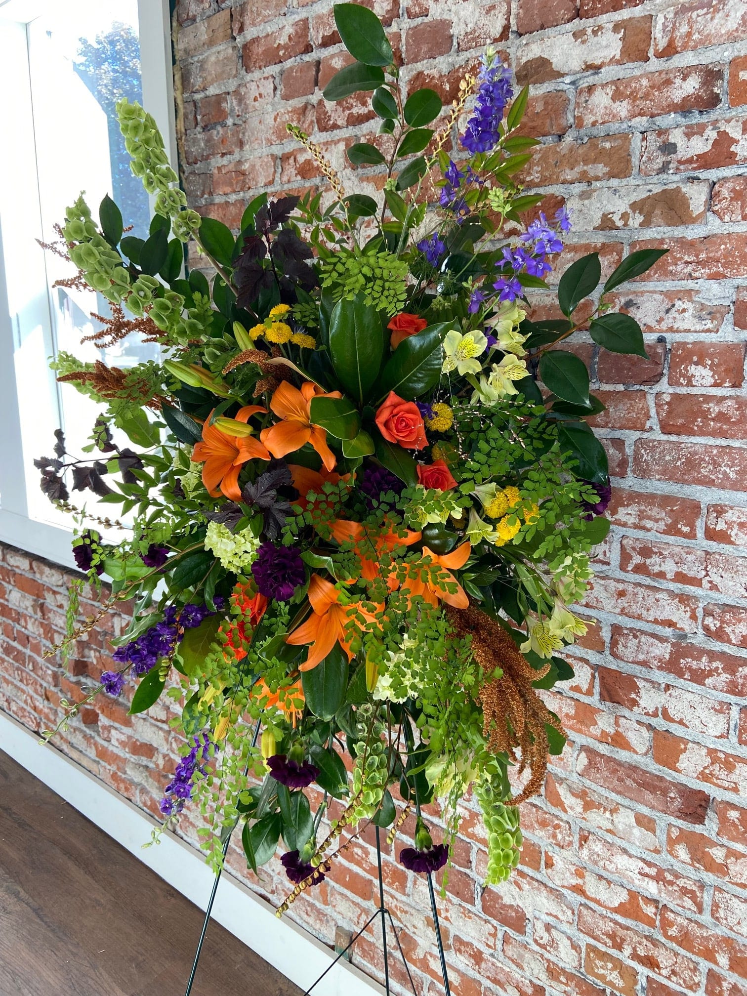 Tall standing spray with orange lilies, roses, purple delphinium and mixed blooms on a metal easel