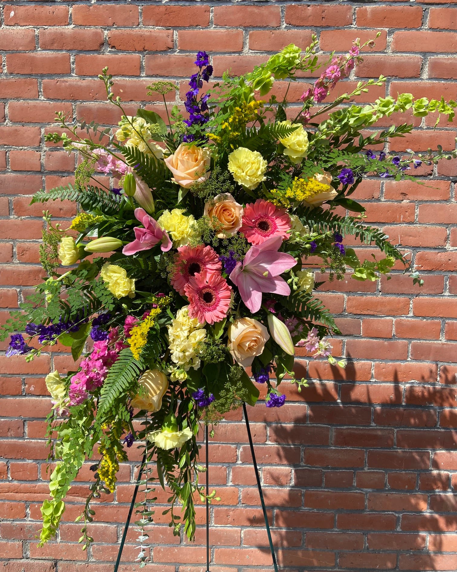 Tall standing spray with pink lilies, peach roses, and yellow flowers on an easel.