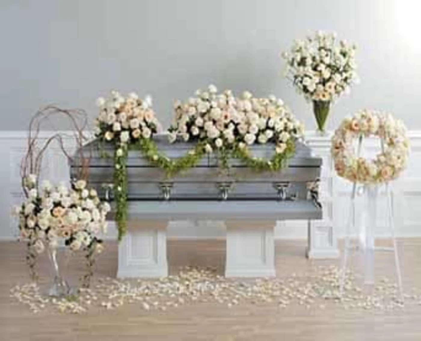 Set of ivory and peach rose funeral arrangements around a gray casket with greenery garlands.