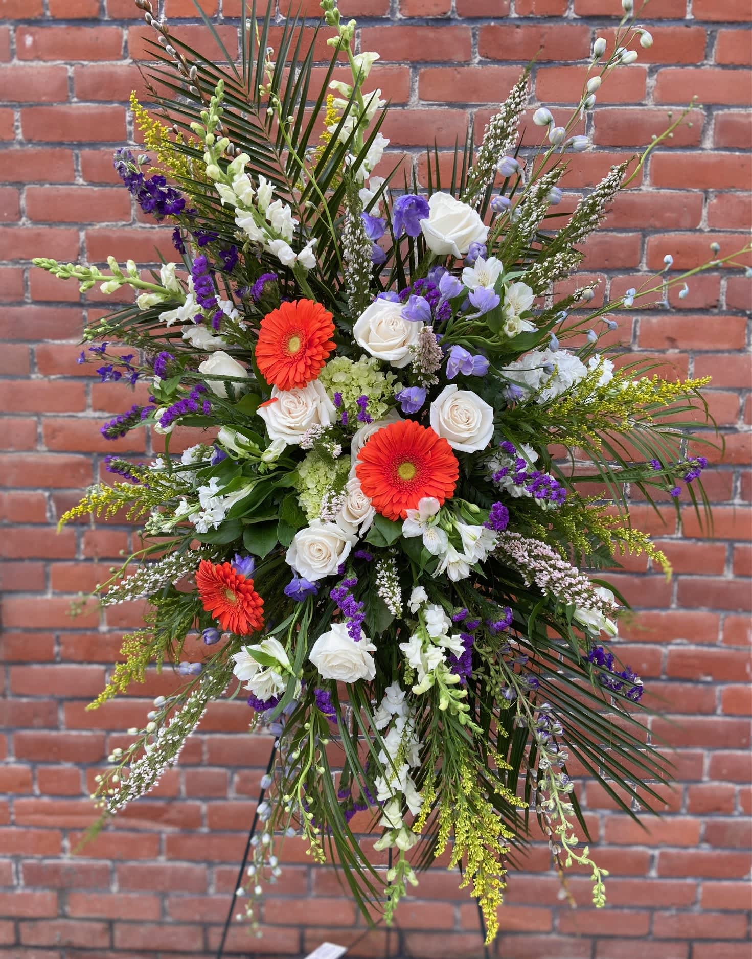 Standing spray of white roses, orange gerbera daisies, and purple blooms with tall palm fronds on an easel.