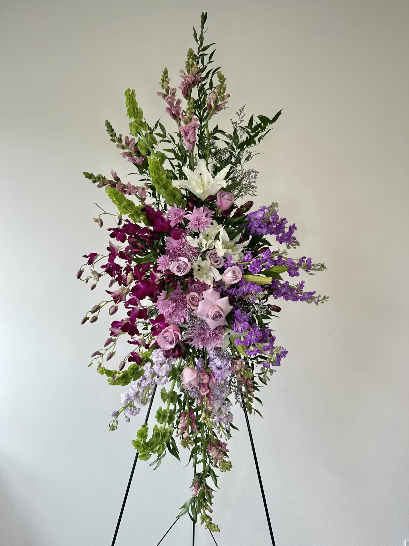 Sacred Garden Spray - A blooming embrace for the tenderhearted. The Sacred Garden Spray from Irvine Village Flowers is a floral whisper of compassion, hope, and grace, crafted to share your deepest sympathy. This lovely lavender spray weaves together a serene tapestry of purple and white blossoms—hydrangeas, orchids, roses, oriental lilies, and more—the standard standing tall at approximately 22" W x 50" H and deluxe at 30" W x 60" H. A one-sided tribute, it’s a gentle way to honor a life with beauty and peace.  Each flower offers its own quiet comfort: lavender hydrangeas spill over with heartfelt abundance and gratitude, their soft clusters a soothing balm. Pure white orchids lift spirits with their elegance, symbolizing innocence and eternal love. Roses in gentle hues breathe reverence and care, while oriental lilies unfurl with a fragrance of hope and serenity. Together, they create a sacred garden in bloom—a tribute that feels like a warm, floral hug for those grieving.  Visit Flower Synergy to send this compassionate spray, delivered fresh to wrap your loved ones in nature’s embrace. It’s a simple, beautiful way to say you’re there, sharing hope and love through every petal.