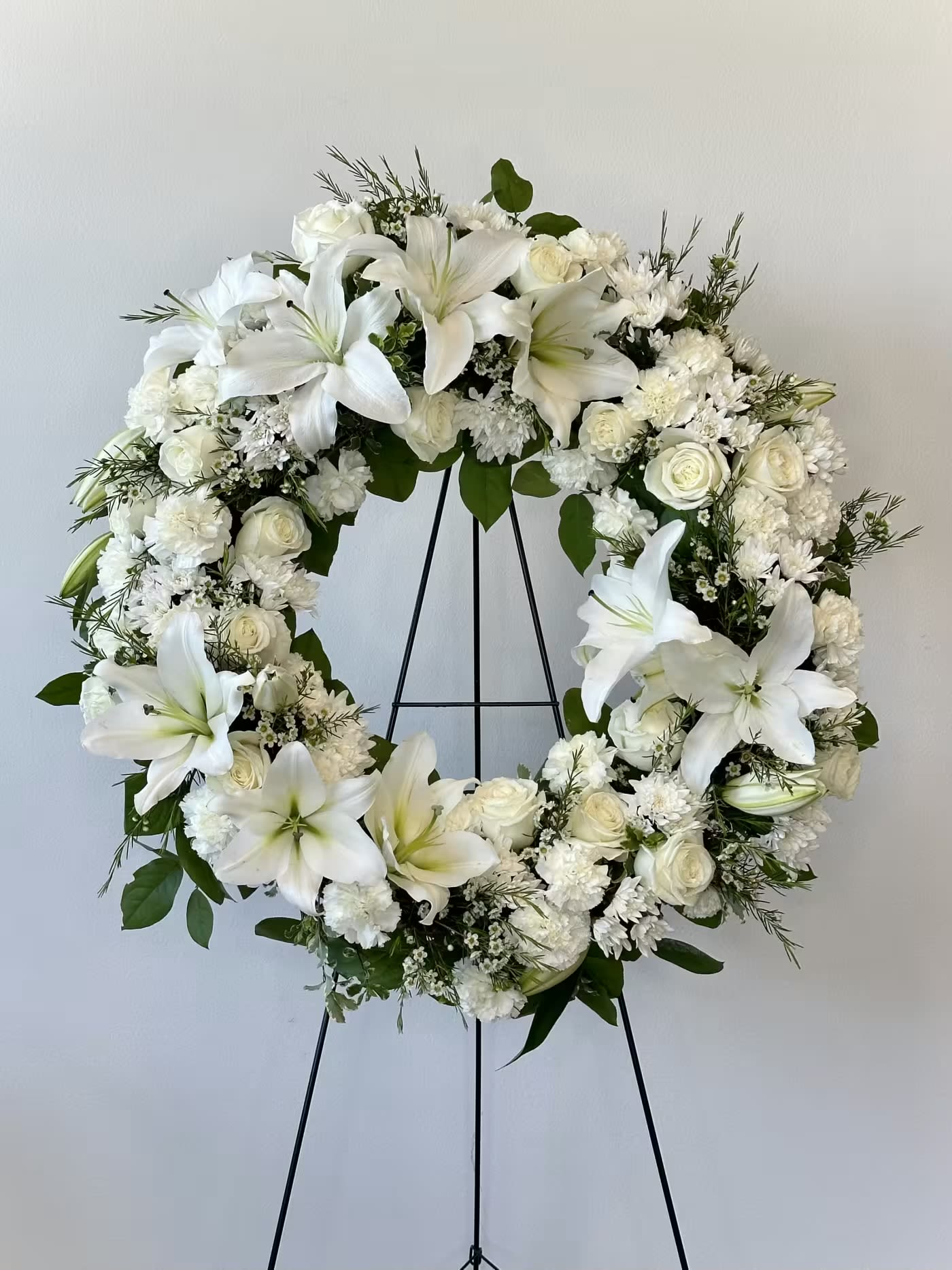 Serenity Wreath - A gentle embrace for tender hearts, the Serenity Wreath from Irvine Village Flowers offers a tranquil ring of fragrant white blossoms, a quiet tribute for any funeral or wake. Delivered on an easel, this classic arrangement whispers sympathy and reverence, crafted with care from fresh roses, Asiatic lilies, carnations, and cushion spray chrysanthemums, each bloom cradled by soft greenery. Measuring 22" W x 22" H, its one-sided grace stands as a thoughtful beacon of peace.  The pure white roses speak of honor, remembrance, and quiet love, their petals a tender solace. Asiatic lilies, radiant and calm, bloom with dignity and hope, easing sorrow with their gentle presence. Carnations offer enduring affection and resilience, while cushion spray chrysanthemums lend a comforting fullness, symbolizing truth and eternal rest. Together, they weave a wreath of serenity, a heartfelt gesture for those in mourning.  In this time of stillness, Flower Synergy is here for you—visit us online or in-store to share this compassionate creation. With our seamless delivery across Irvine, your tribute arrives fresh, a delicate balm to honor and console.